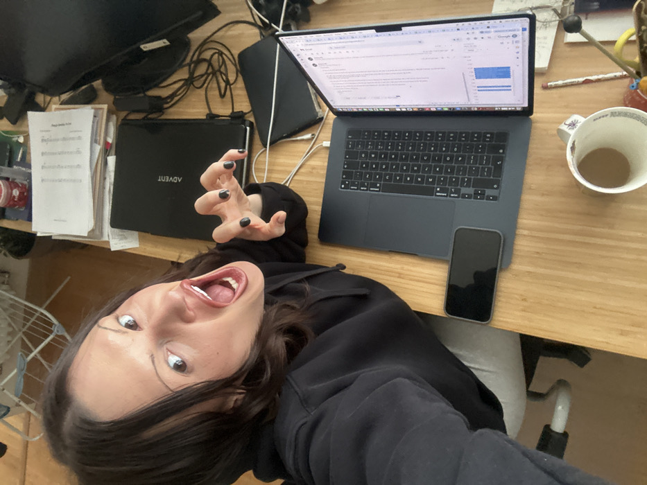 This image depicts a single female subject positioned at a wooden desk in an indoor setting, identified as a location within Tunbridge Wells, United Kingdom. The female, with dark hair, is wearing a black hooded sweatshirt and light-colored trousers, visible from an overhead perspective. Her face is directed towards the camera with an exaggerated expression: eyes wide open, eyebrows raised, and mouth wide open, revealing the oral cavity. Her right hand is raised, fingers curled into a claw-like gesture, with black nail polish visible.
The desk surface, made of light-colored wood, holds several items. To the right of the female, a silver-gray MacBook laptop is open with its screen vertical, displaying what appears to be a spreadsheet or document with text and blue bar graphs. A black smartphone with a dark screen is positioned horizontally to the left of the MacBook. Further to the right and behind the MacBook, a closed black laptop with the "ADVENT" logo is visible, alongside a pile of documents and loose papers. Numerous tangled white and black cables are also present on the desk, connecting various electronic devices. A white mug containing a light-colored liquid, possibly coffee or tea, is situated in the lower right corner of the frame. In the background, behind the desk, a white clothes drying rack and a green potted plant are partially visible, indicating a domestic or office environment.