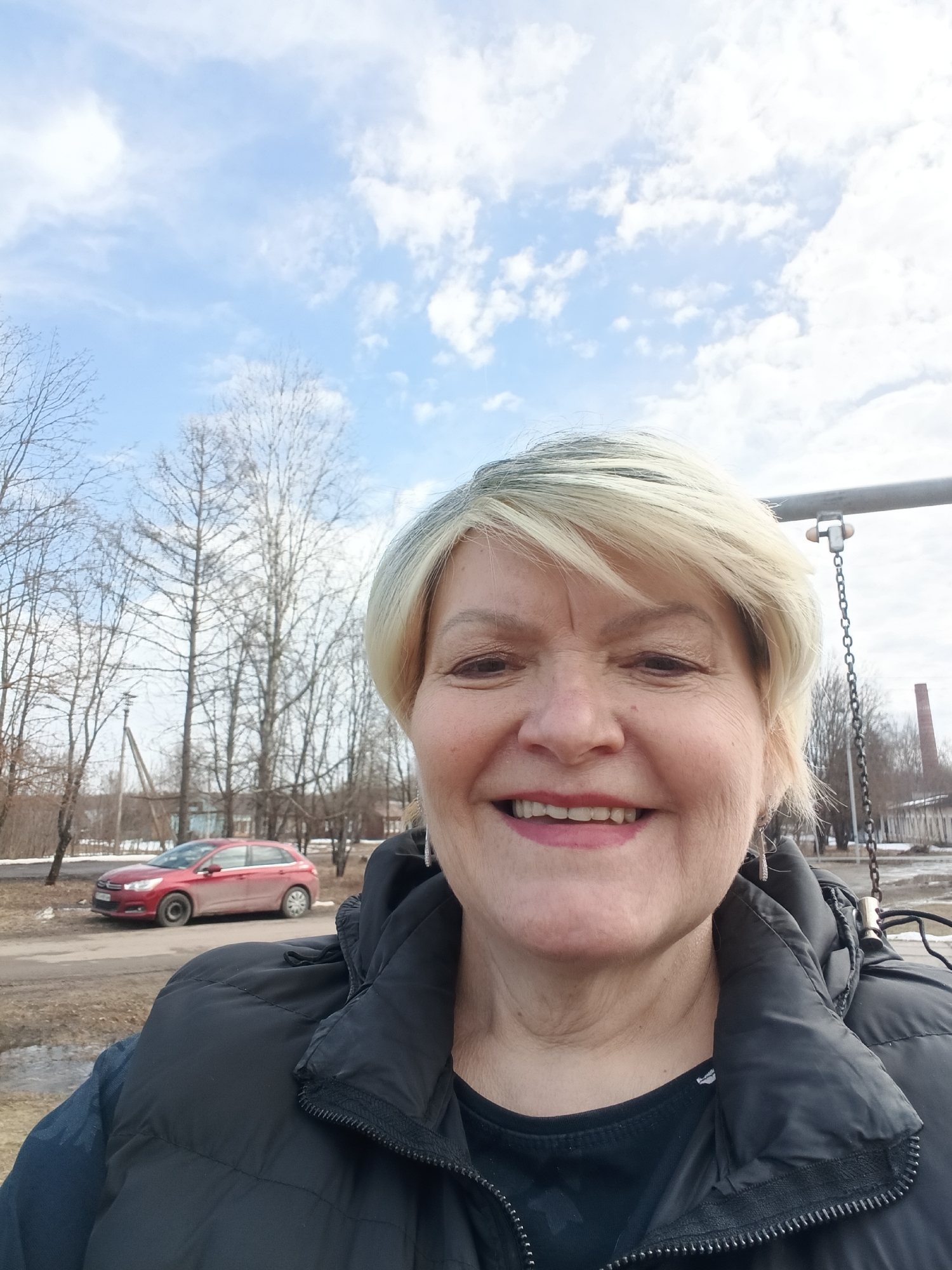 The image is a selfie of a smiling mature woman with blonde hair. She's wearing a black puffer vest and a dark shirt. In the background, a red car is parked near bare trees, indicating a season like early spring or late autumn. The sky is light blue with scattered white clouds. There is a swing set structure visible to the right of the woman, and in the distance, some buildings and what appears to be a brick chimney are visible. Based on the location in the city of Красный Маяк, Russia, the environment appears to be a residential or park area.