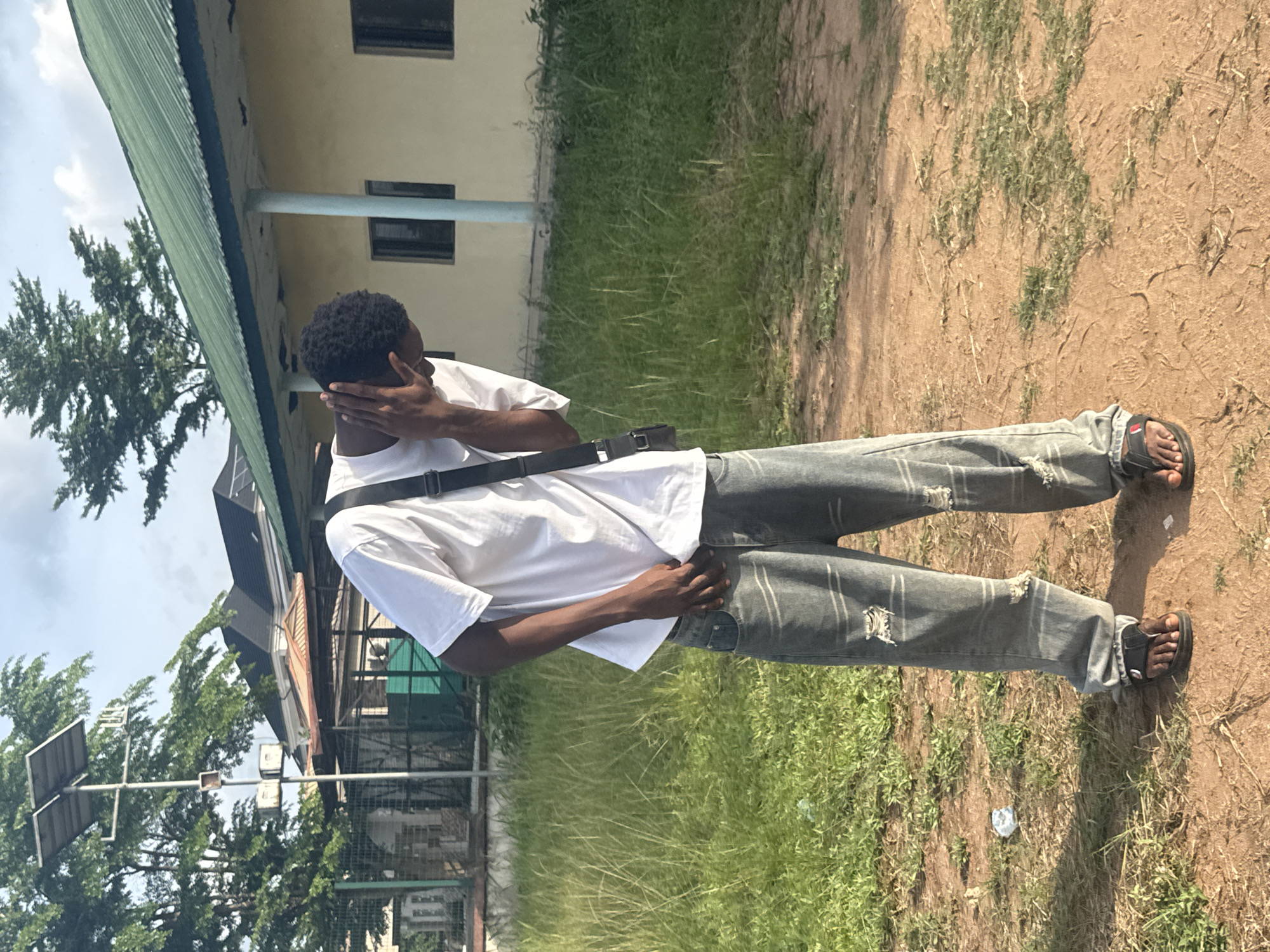 A person with dark skin is standing on an unpaved patch of land with some grass, likely a roadside in Ihiagwa, Nigeria. They are wearing a white t-shirt, gray ripped jeans, and black sandals, and have a black strap across their chest. The person is holding their head with their hand. Behind the person is a building with a green corrugated roof and a light-colored facade. There are trees nearby. The sky is visible in the background.