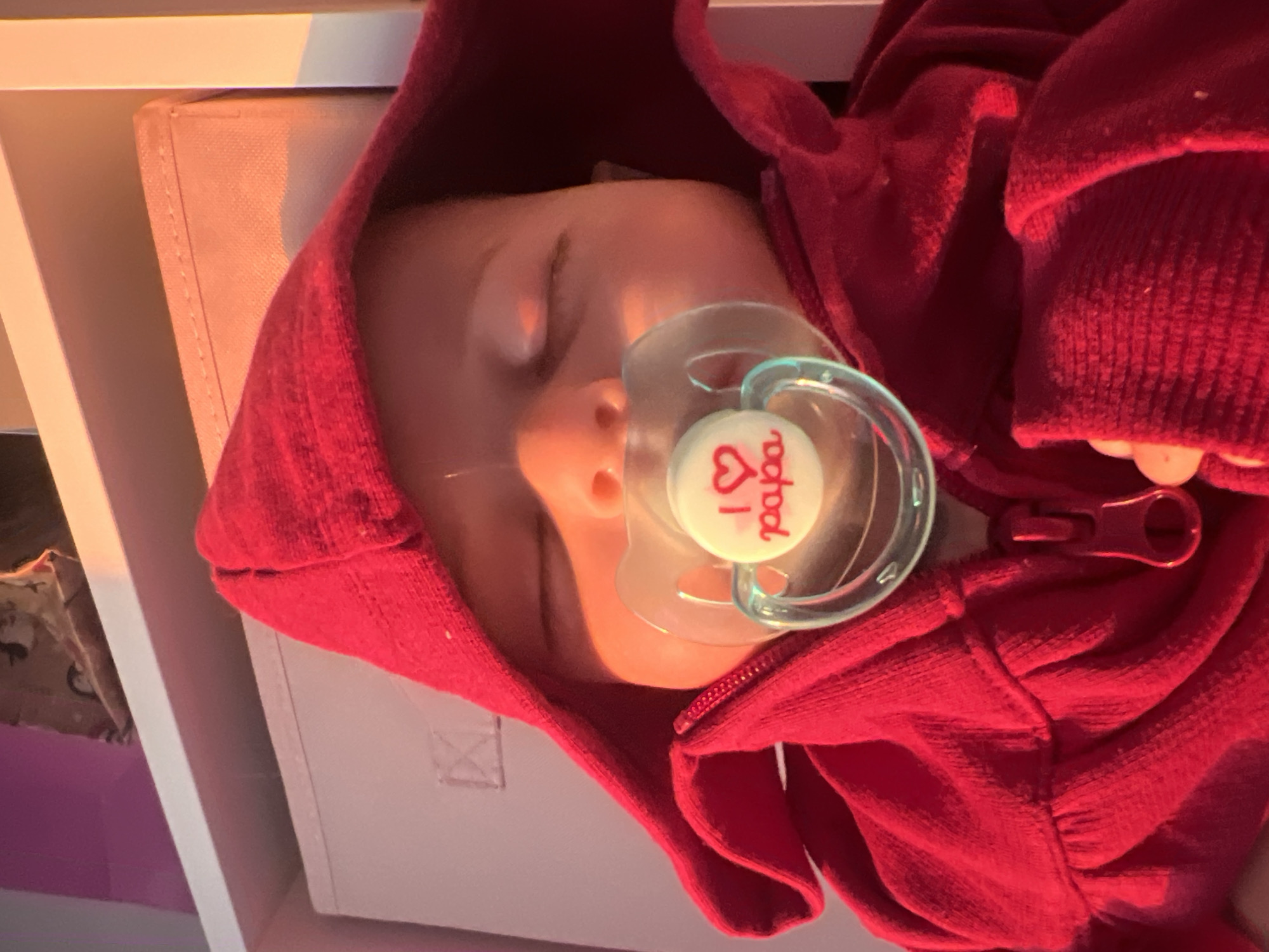 The image shows a close-up of a baby sleeping, wearing a red hooded garment and a pacifier. The pacifier has a white shield with a red heart and the word "papa" written in red. The baby is lying on its back, with its eyes closed. The background appears to be a shelf in what might be a child's room, with some storage boxes visible. The lighting suggests it might be late afternoon or early evening. The image was taken in Votkinsk, Russia.