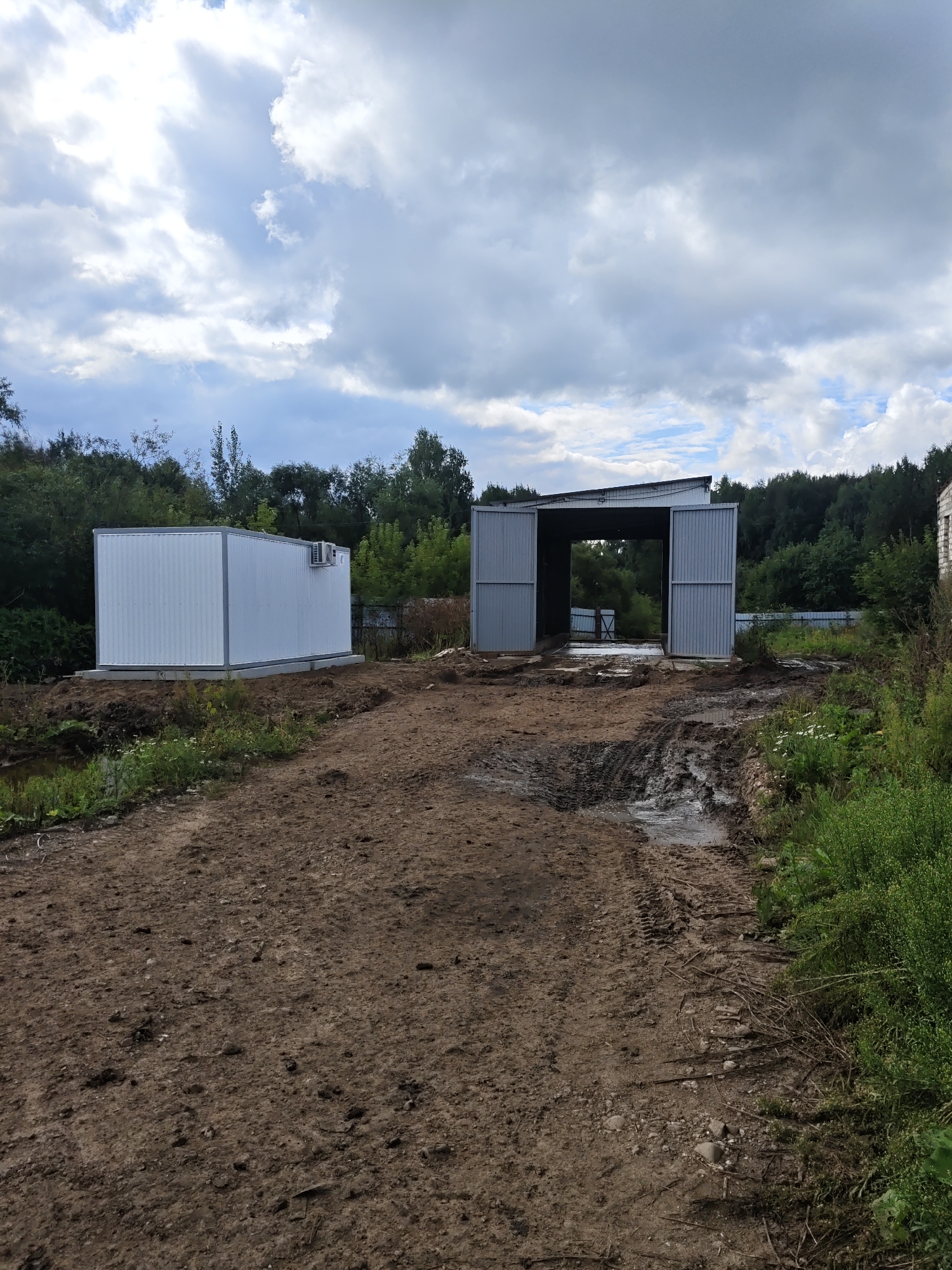 In Платошино, Russia, an outdoor scene depicts the construction or development of two modular metal structures amidst a rural or semi-rural environment. In the foreground, a dirt path with visible tire tracks and puddles of water suggests recent activity or rainfall, indicating a muddy terrain. To the left, a rectangular white modular building, possibly a prefabricated container or office, is situated on a concrete foundation, with an air conditioning unit mounted on its exterior. Further to the right, a larger, open-sided metal structure, resembling a garage or workshop with a gabled roof, stands with its wide doors open. This structure also appears to be on a prepared base, facing a muddy area. The background is dominated by dense green trees and foliage, typical of a wooded or forested area. The sky above is overcast with dramatic clouds, suggesting a dynamic weather pattern. The overall impression is one of an ongoing construction or agricultural project in a natural setting.