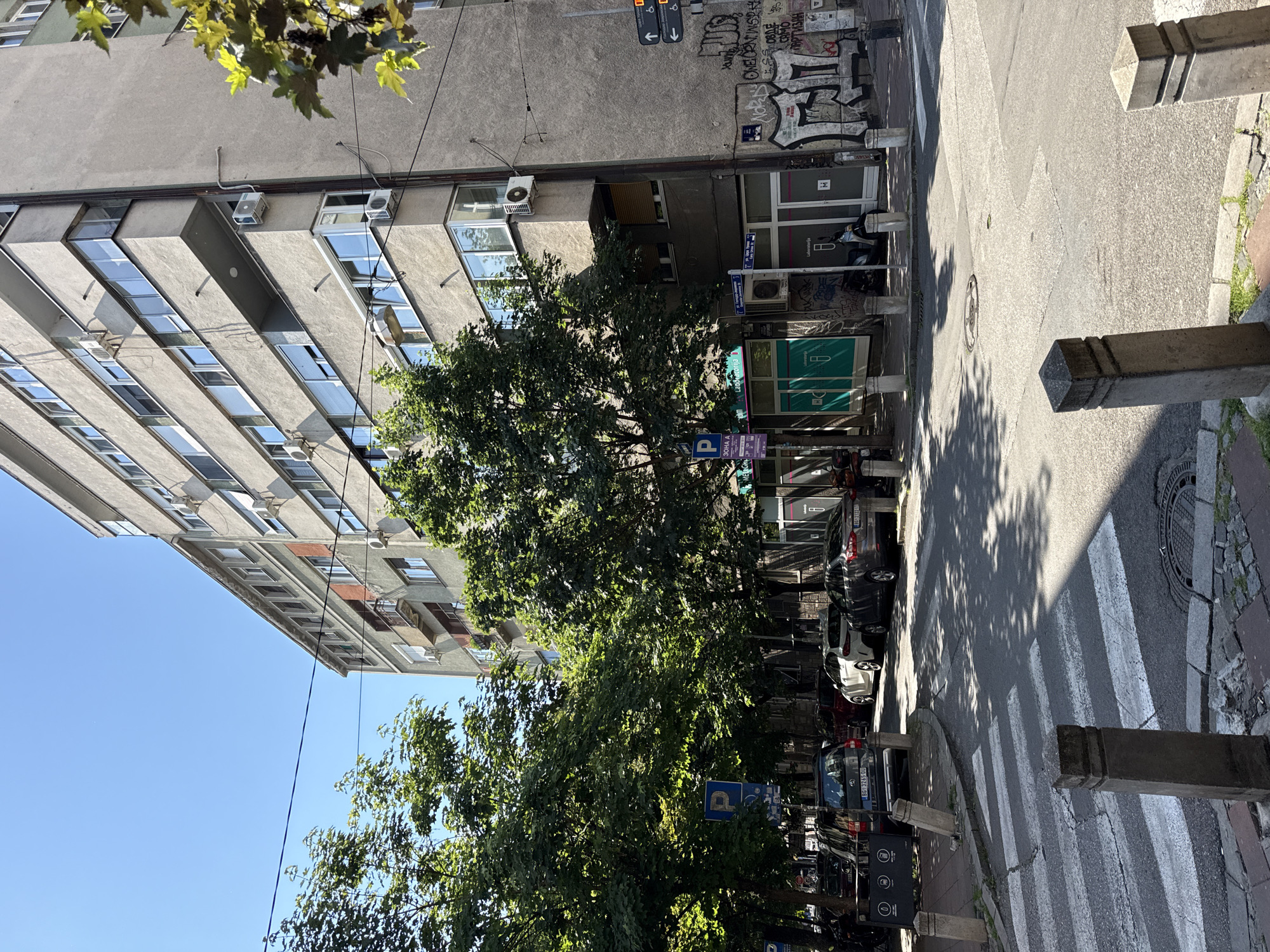 This is an eye-level, daytime shot of a street scene in Belgrade, Serbia. The camera is positioned on a street corner, near a crosswalk.
The street is lined with parked cars. A tall, gray, multi-story building rises on the left side of the frame, with rows of windows and numerous air conditioning units attached to the exterior. Trees with dense green foliage dominate the foreground and obscure portions of the building and street.
Street signs and parking signs are visible amidst the trees and parked cars. Graffiti adorns the lower portion of the building on the right side of the frame. The crosswalk is marked with white stripes and has concrete bollards on either side. The road surface shows signs of wear and repair, including cracks and patches. The sky visible above the building is a clear blue. There are no people visible in the scene.