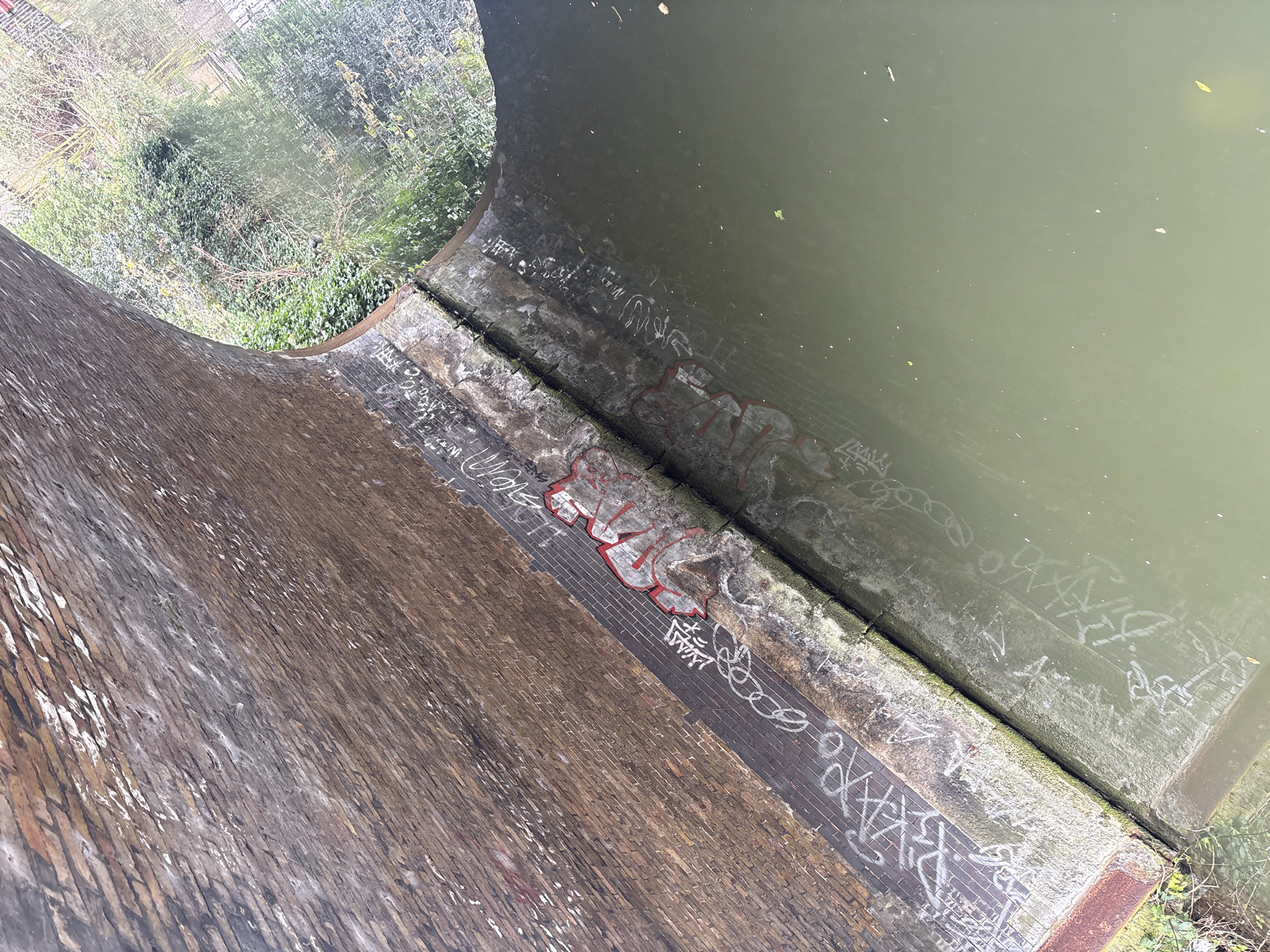An aerial view shows a brick structure bordering a body of water. The bricks are a mottled brown, stained with moisture and marked with graffiti. Some of the graffiti is in red and white. The water is a murky green. A portion of the brick structure arches, revealing trees and vegetation behind it. The scene appears to be a canal or waterway structure, likely located in London, United Kingdom.