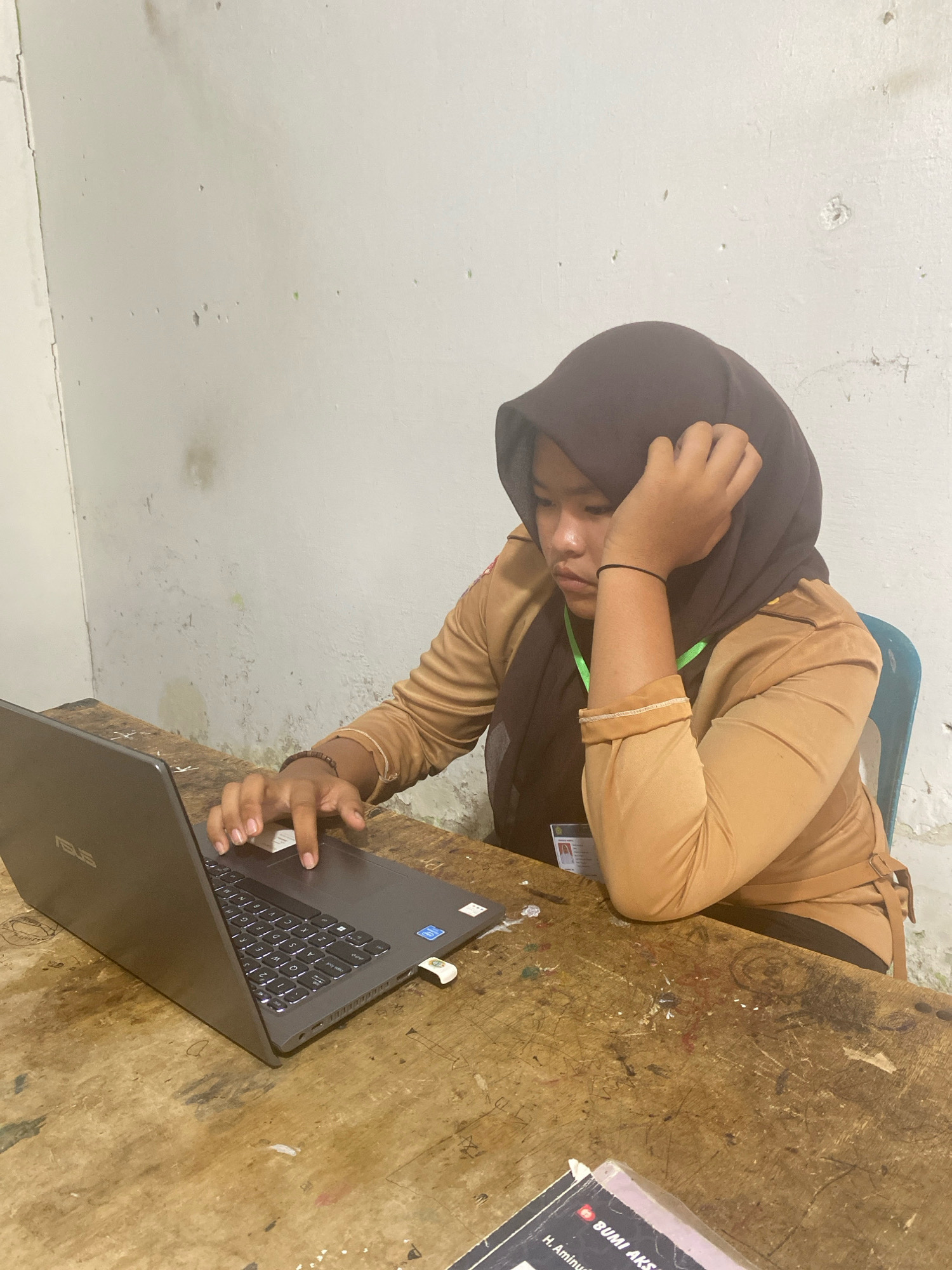 A young Indonesian woman, wearing a brown uniform and a dark hijab, is seated at a wooden desk, looking at a laptop. Her right hand rests on her chin, and her left hand is on the laptop's touchpad. The desk is worn and marked with graffiti. The background is a textured, light-colored wall with some imperfections. A blue chair is visible to the right. The scene appears to be indoors, possibly in a classroom or office setting in Karang Gading, Indonesia.