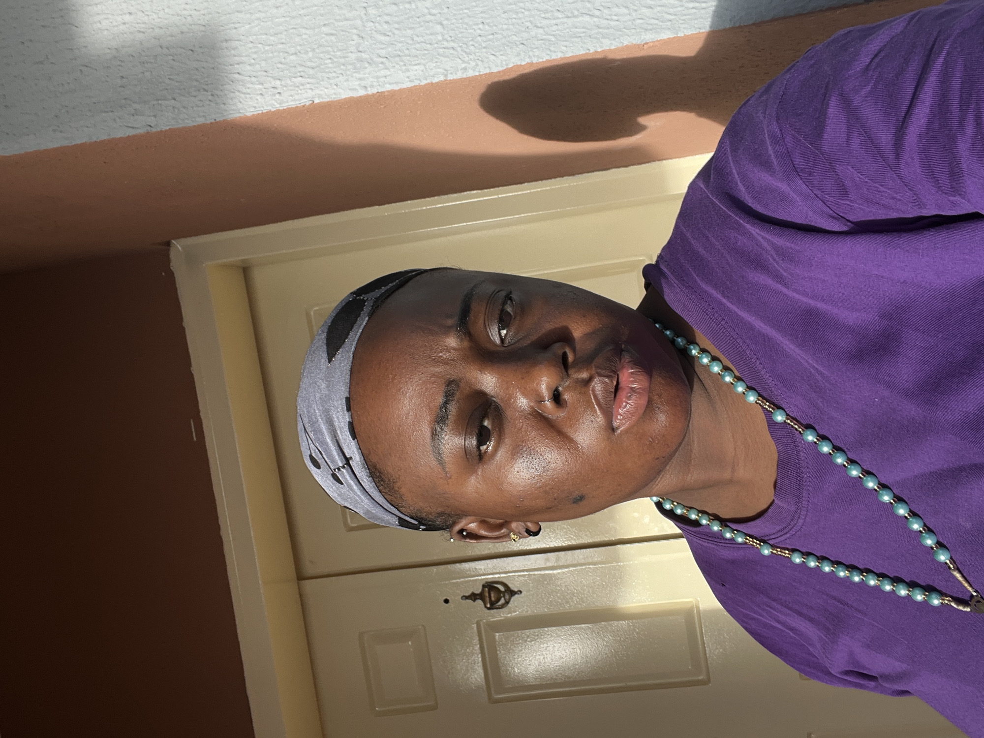 An outdoor portrait, rotated 90 degrees counter-clockwise, features a person with dark skin in Jos, Nigeria, looking directly at the camera with a neutral and observant expression. The individual wears a long-sleeved purple t-shirt and a grey head covering with a black pattern. A light blue beaded necklace with gold links is visible, draped over their shirt. Notable details include a nose piercing, a black hoop earring, and a gold stud earring.
The setting appears to be an exterior space or a very brightly lit area, possibly a porch or entrance. Behind the person is a cream-colored door, featuring recessed panels and a metallic door knocker or handle. Above the door, there's a horizontal strip of light brown painted wall, topped by a textured white wall or ceiling. Strong, direct sunlight illuminates the scene, casting bright highlights on the person's face and shirt, and creating distinct, dark shadows on the brown wall, indicating a sunny day, likely in the morning or afternoon. The bright lighting and sharp shadows suggest clear weather. No visible text is present in the image.
