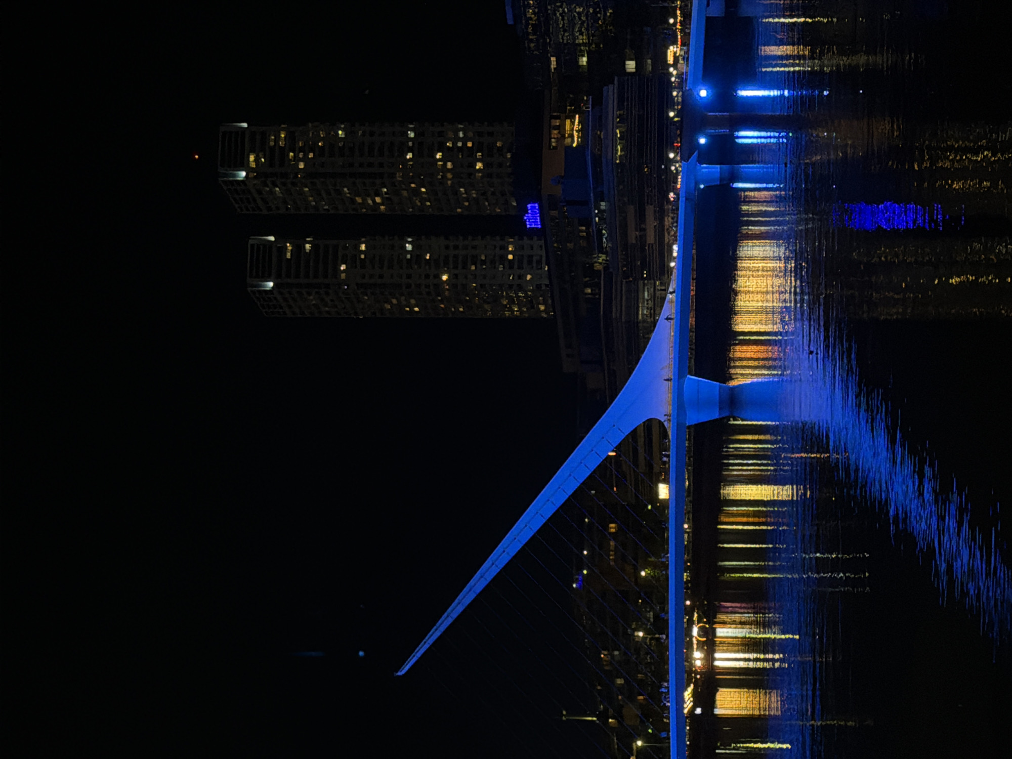 Night view in Buenos Aires, Argentina. A blue illuminated pedestrian bridge, the Puente de la Mujer, crosses a body of water reflecting light from buildings and other structures. A tall building with numerous illuminated windows is seen on the other side, bearing the lit-up name of Hilton. The sky is dark.