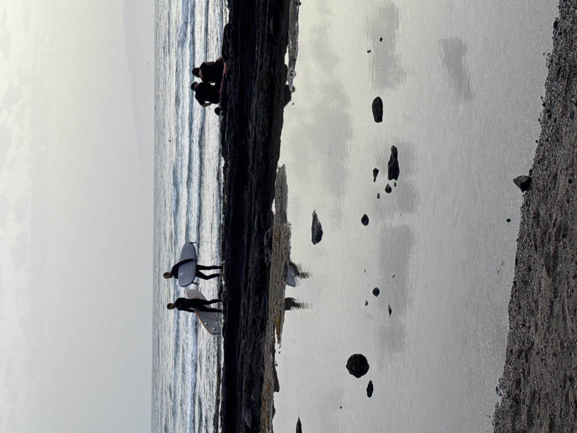 The image shows a rocky coastline with several people. In the foreground, a shallow tide pool reflects the sky and the surrounding rocks. Two surfers in wetsuits are walking on the dark rocks near the water's edge, each carrying a surfboard. Further back, two other individuals are seated on the rocks, facing the ocean. The ocean itself is calm with gentle waves. In the distance, a faint outline of land is visible on the horizon. The overall lighting suggests it is early morning or late evening. The scene is consistent with a location along the coast of Playa De Las Américas, Spain, known for its volcanic rock formations and surf spots.