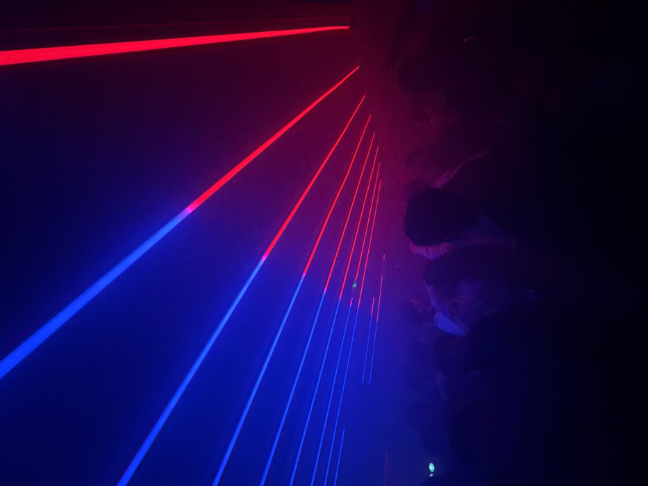 The image shows a dark room filled with vibrant red and blue laser beams. The lasers run diagonally across the frame, creating a dynamic and futuristic atmosphere. On the right, there are dark, blurred shapes that look like people standing in the room, barely visible in the dim lighting. The scene evokes a sense of energy and excitement.