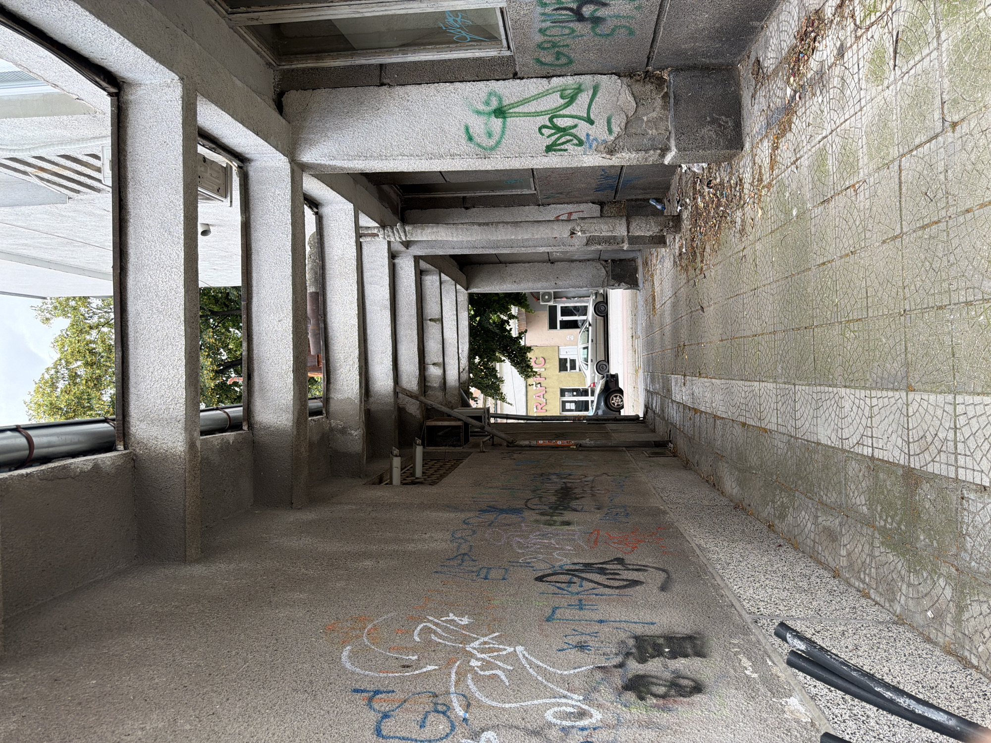 The image shows an exterior view of a covered walkway or alleyway. The walkway is paved with patterned tiles and is flanked by walls on the left and a building with a series of concrete columns and beams on the right. The walls are covered with graffiti. Debris and leaves are scattered on the ground along the right side. In the background, a car is parked in front of a building with a yellow facade and the word "TRAFFIC" is visible on a sign. Trees are visible above the buildings. The overall scene suggests an urban environment, possibly a side street or an area between buildings. The specific location in Pazardzhik, Bulgaria, is identified through the graffiti style and architectural elements common in the region.