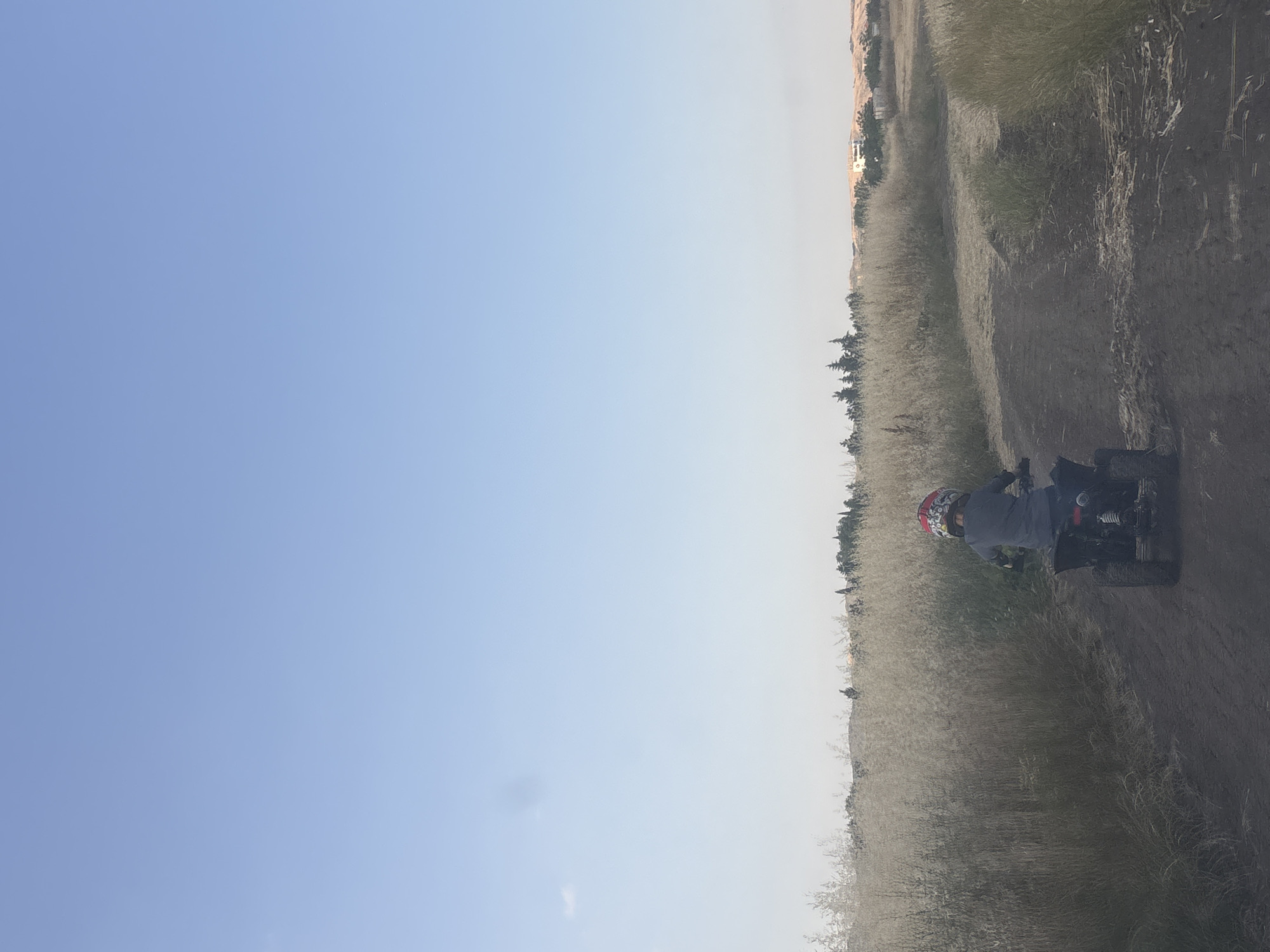 The image shows a young child wearing a helmet riding a black ATV on a dirt path. The child is facing away from the camera, toward the right side of the frame. The path is surrounded by dry, tall grass. In the background, there are trees and a few buildings, suggesting a rural or semi-rural setting. The sky is clear and light blue. The scene appears to be in a dry, possibly summery environment. This image is unlikely to be in the city of Burgas, Bulgaria, as the terrain and vegetation do not strongly suggest a Bulgarian coastal landscape.