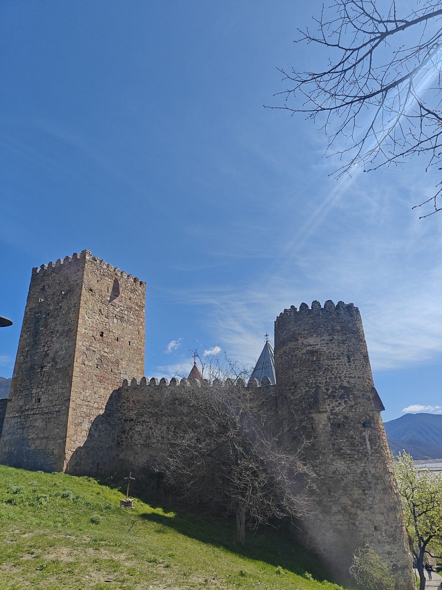 An outdoor, low-angle shot captures a historic stone fortress or monastery complex under a bright blue sky. Two prominent stone towers, connected by a crenellated wall, dominate the scene. The left tower is rectangular and taller, featuring multiple small openings, while the right tower is rounded. A conical, possibly metallic, roof with a cross on top is visible behind the wall, suggesting a church or central building within the complex.
The structures are built from rough-hewn stones, showing signs of age. The complex is situated on a grassy slope, with a simple wooden cross planted in the ground near the base of the left tower. Bare tree branches, some with tiny buds, frame the upper right corner and appear along the wall, indicating early spring. In the distant background, rolling mountains can be seen under the clear sky, with what might be a valley or body of water further beyond. Two faint human figures are visible walking on a path to the far right, near a tree with light green foliage.
The time of day is bright daylight, likely midday or early afternoon, with strong sunlight visible as a lens flare and rays emanating from the upper right portion of the sky. The sky is a vibrant blue with sparse, wispy clouds. No discernible text is visible in the image. The overall impression is one of historical significance and serene nature.