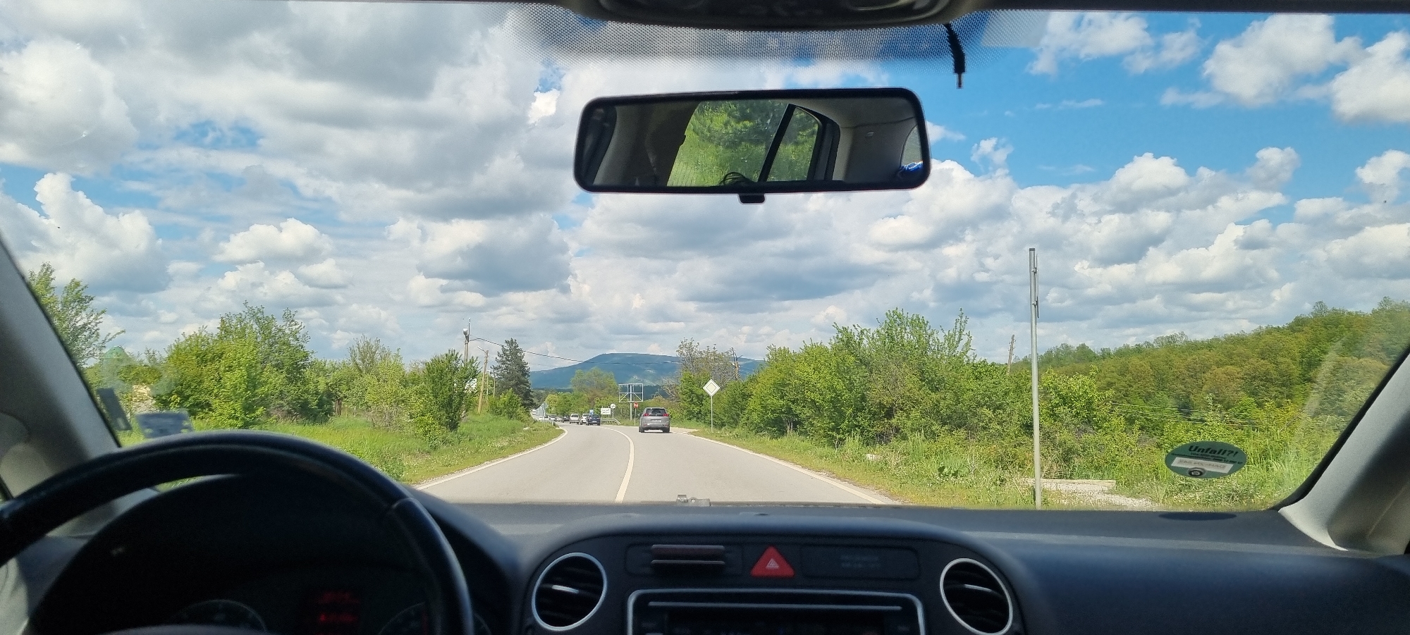 The image depicts a view from inside a car, looking forward onto a winding road outside Blagovo, Bulgaria. A grey vehicle is directly ahead on the road, with other cars appearing in the distance. Lush green vegetation lines both sides of the road. The sky above is blue with puffy white clouds. The dashboard of the car is visible. A rearview mirror is positioned at the top of the windshield. A utility pole stands by the side of the road. A sticker is seen attached to the windshield on the passenger side.