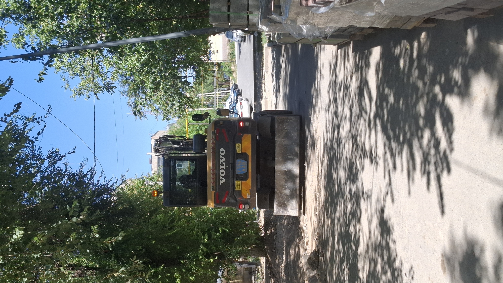 An excavator with the Volvo logo is parked on a paved surface, partially shaded by trees. The excavator is yellow and black with a large bucket attachment at the rear. It has orange hazard lights visible on the top. The scene appears to be in an urban or suburban area, as indicated by a building visible in the background and parked cars further down the road. The ground is a mix of asphalt and dirt, with some construction materials, possibly paving stones, stacked on the right side of the image. The sky is clear and blue. The overall impression is one of a construction or maintenance site within a city. This scene is located on the street of Akimat, in the city of Shymkent, Kazakhstan.