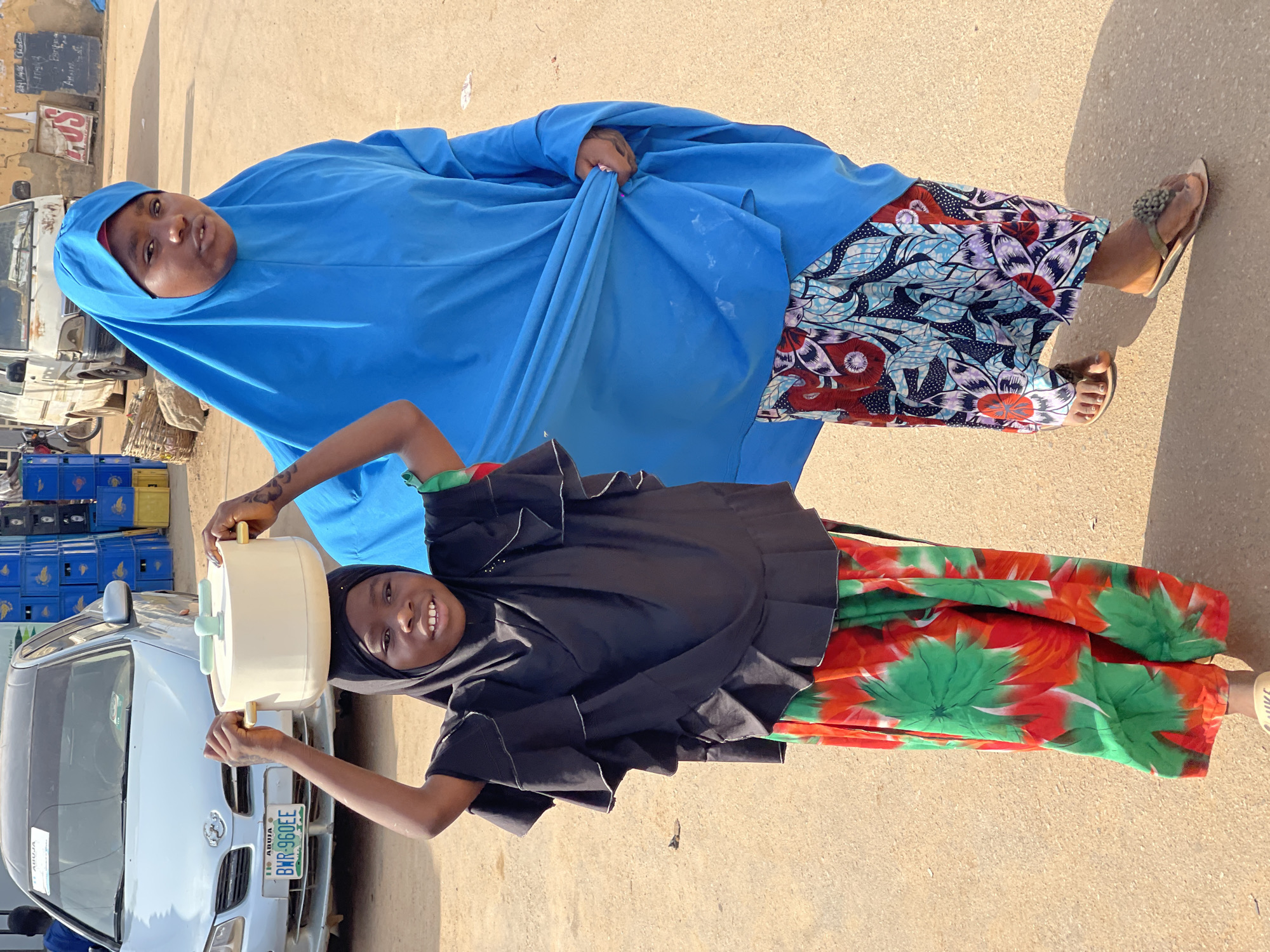 The image features two females, an adult and a child, standing outdoors on a sunny day in what appears to be an urban or market setting in Kuchingoro, Nigeria. The ground is a light-colored paved surface, likely asphalt or concrete, with strong shadows indicating bright daylight.
The adult, positioned in the background, wears a vivid blue hijab covering her head and body, paired with a colorful patterned skirt or dress featuring blue, red, purple, and white floral or leaf designs. She has light-colored sandals with green beaded embellishments.
In the foreground, a younger female is smiling cheerfully at the camera. She wears a black hijab and a brightly patterned skirt with prominent red and green floral or leaf motifs. She expertly balances a light cream-colored container on her head, steadying it with both hands on its gold-colored side handles. The container also has a light blue-green handle or knob on top. A tattoo is visible on her right forearm.
The background includes a silver Toyota sedan to the left, displaying a Nigerian license plate that reads "ABUJA BWR-960EE". Behind the adult, an older, light-colored truck or van is parked. Stacks of blue crates are visible near the sedan. On a wall behind the adult, there's a partially visible red and white sign with "LOS" printed on it, along with other faint text.