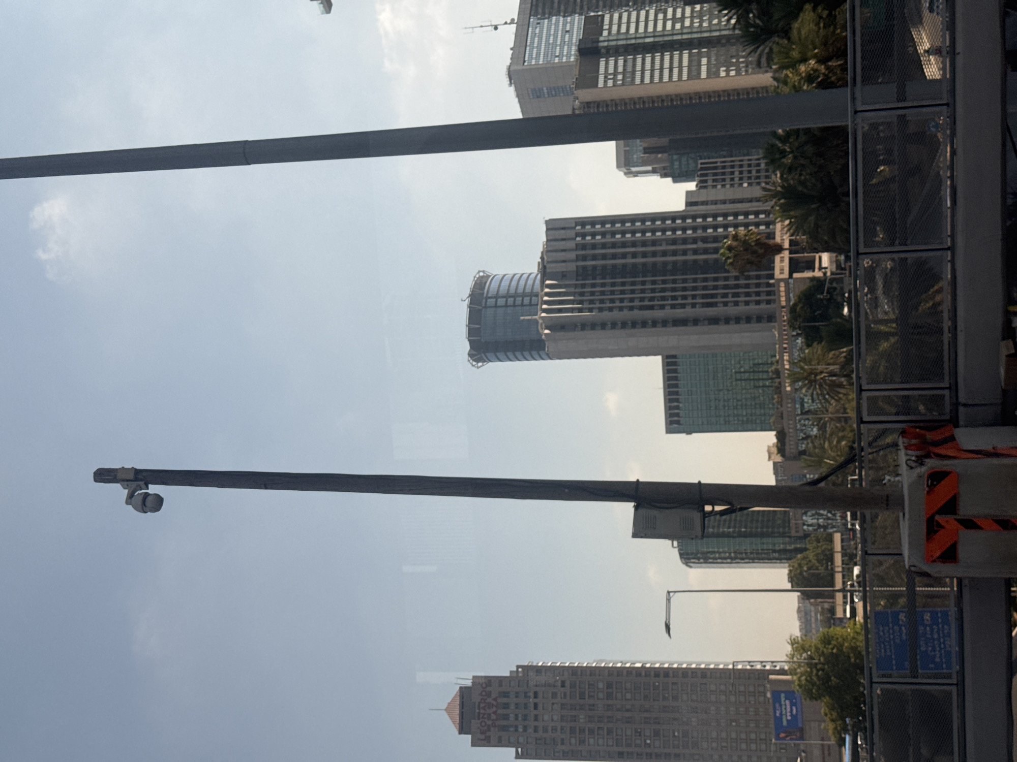 The image captures a view of the Tel Aviv skyline, likely from an elevated position such as a bridge or overpass, with a clear sky and scattered clouds. The foreground features a metal fence with mesh panels and reflective orange and black markers on a concrete post, indicating a safety or traffic barrier. A security camera is mounted on a tall wooden pole in the mid-ground.
Dominating the scene are several modern skyscrapers of varying heights and architectural designs. On the left, a tall, rectangular building with a reddish-orange pyramid-shaped roof is prominently visible. Text on this building appears to read "LEONARDO PLAZA". To its right, a distinctive cylindrical skyscraper with a dark, textured facade and a circular structure at its top stands out. Further to the right, a cluster of other high-rise buildings, some with glass facades and detailed architectural features, complete the urban landscape. Palm trees and other greenery are visible between and around the buildings, suggesting a well-developed urban area with landscaping. Traffic and street lights are discernible in the distance, indicating an active city environment. The overall impression is that of a modern, bustling city on a bright day.