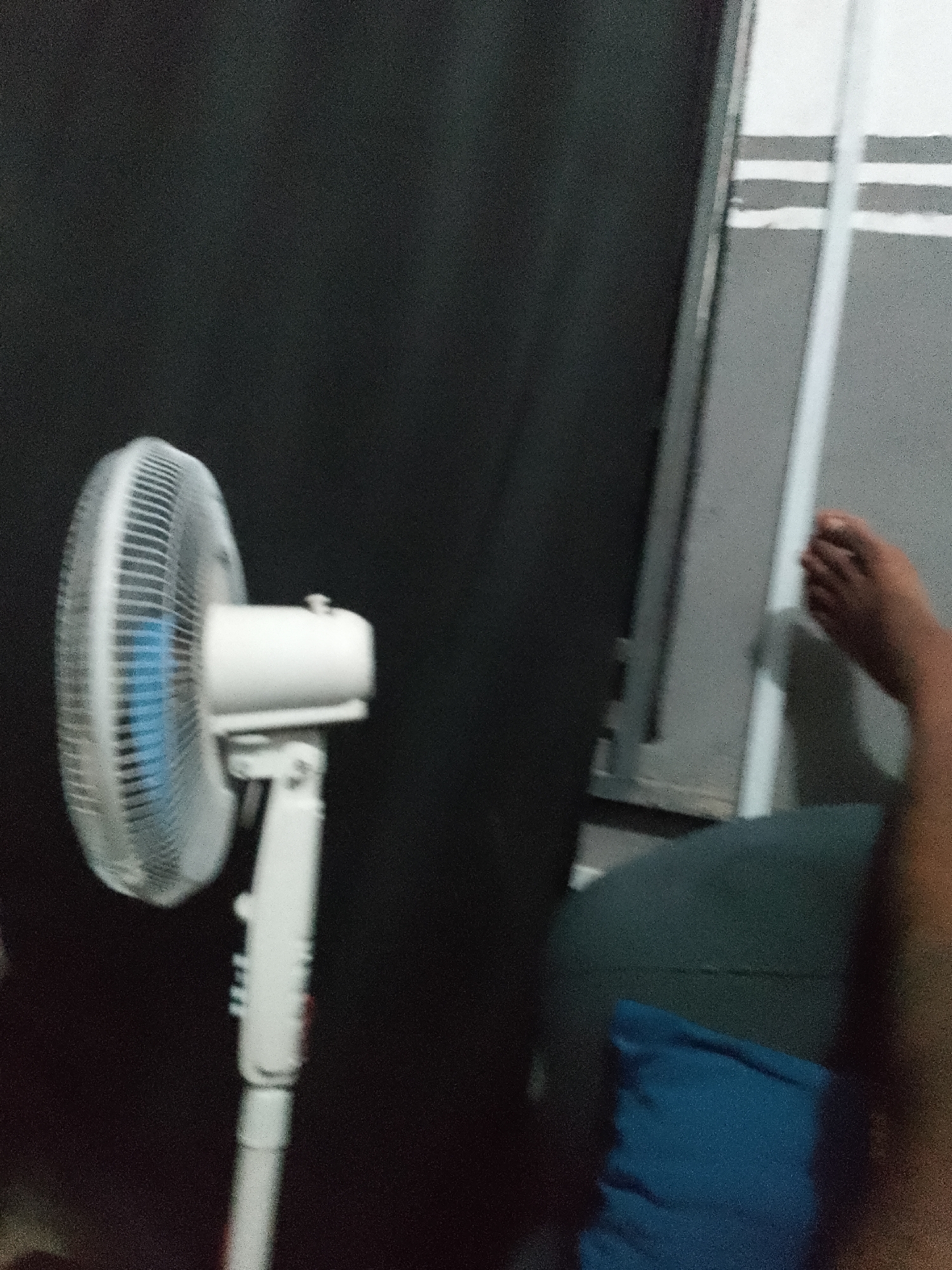 An indoor, eye-level, medium shot captures a white oscillating fan in the foreground, positioned to the left of the frame. The fan, with its blades blurred by motion and a hint of blue visible, is mounted on a white pole. Behind the fan, a dark, textured surface dominates the background, possibly a curtain or a wall. To the right of the fan, a gray, slatted surface with white stripes runs vertically, resembling a wall or a ventilation grate. A person's bare foot and lower leg are visible in the upper right quadrant of the image, resting on a dark surface, possibly a sofa or chair, with a folded blue pillow in front of it. The scene appears to be set in an urban environment, though specific details about the city and its location are not discernible from the image.