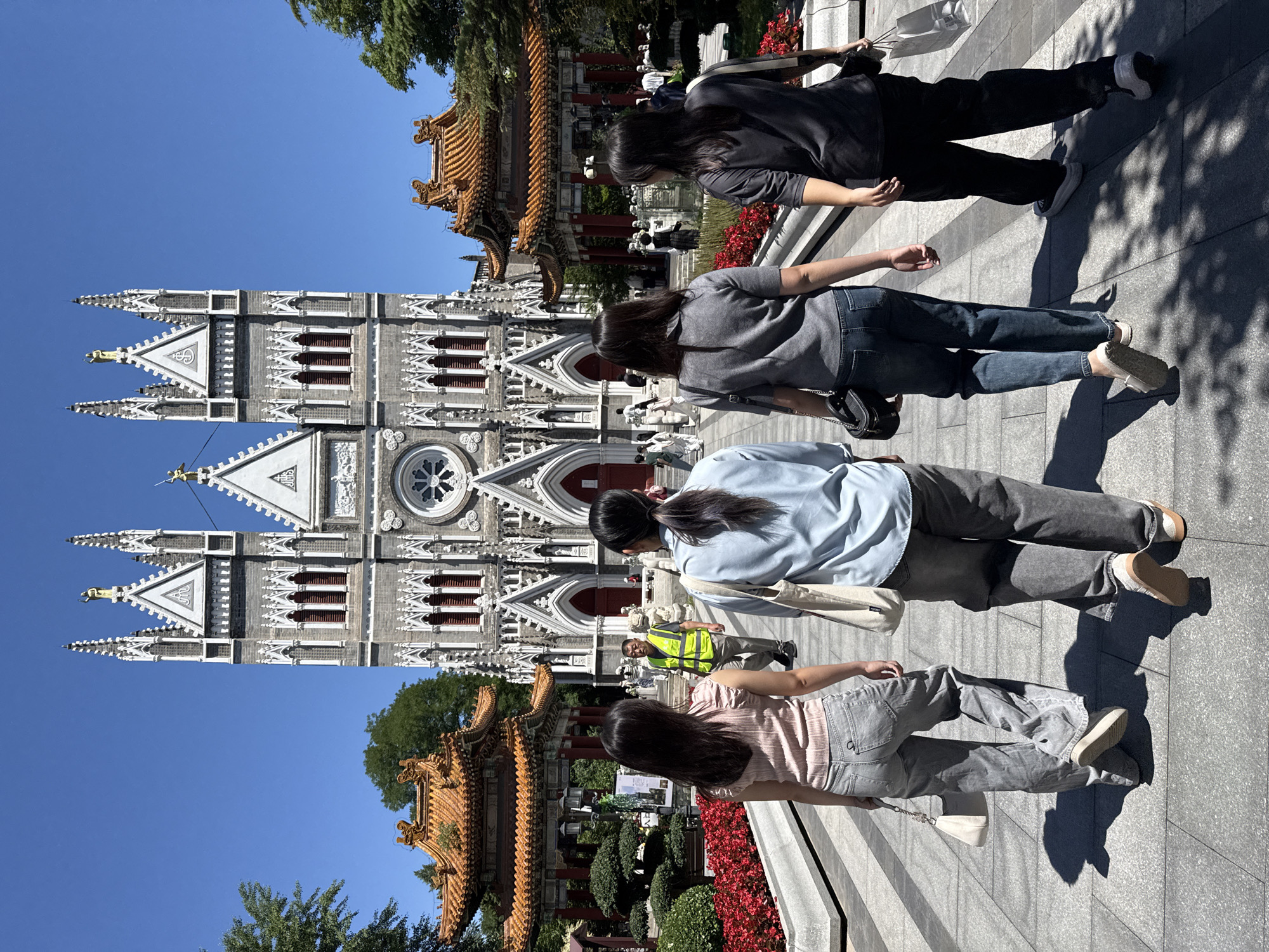 This image presents a detailed view of the Xishiku Church (North Church) and its surrounding grounds in Xicheng District, Beijing, China, under a clear, bright blue sky.
In the foreground, four women are observed from behind, walking along a wide, light-gray paved pathway towards the church. From left to right: the first woman wears a light pink ruffled short-sleeved top, light gray wide-leg jeans, and carries a small white shoulder bag; the second woman wears a light blue long-sleeved shirt, dark gray wide-leg pants, and carries a light-colored tote bag over her left shoulder; the third woman wears a gray short-sleeved top, blue jeans, and a black shoulder bag; and the fourth woman wears a dark gray short-sleeved top, dark pants, and holds a white shopping bag in her left hand. All four women cast distinct, elongated shadows on the pavement, indicating strong sunlight.
The paved pathway is bordered by raised planters filled with dense, vibrant red flowering plants, possibly petunias or impatiens. To the left and partially visible on the right in the midground, traditional Chinese-style pavilions with multi-tiered orange tiled roofs and red pillars stand amidst manicured green foliage, showcasing a blend of cultural architecture. Several other people are dispersed along the path and near the church entrance; notably, a man in a bright yellow reflective vest and light-colored pants is seen walking towards the camera, slightly off-center to the left.
The dominant structure in the background is the Xishiku Church, a large Gothic Revival-style Roman Catholic cathedral. It is constructed from light gray stone or brick and features two prominent bell towers with spires topped by statues, including a golden figure on the left spire. The facade is adorned with intricate white Gothic ornamentation, pointed arched windows fitted with red louvers, and a large circular rose window centrally located above the main entrance. A triangular pediment featuring the "IHS" monogram is visible above the rose window. Lush green trees flank the church and pavilions, providing a natural frame against the cloudless blue sky.