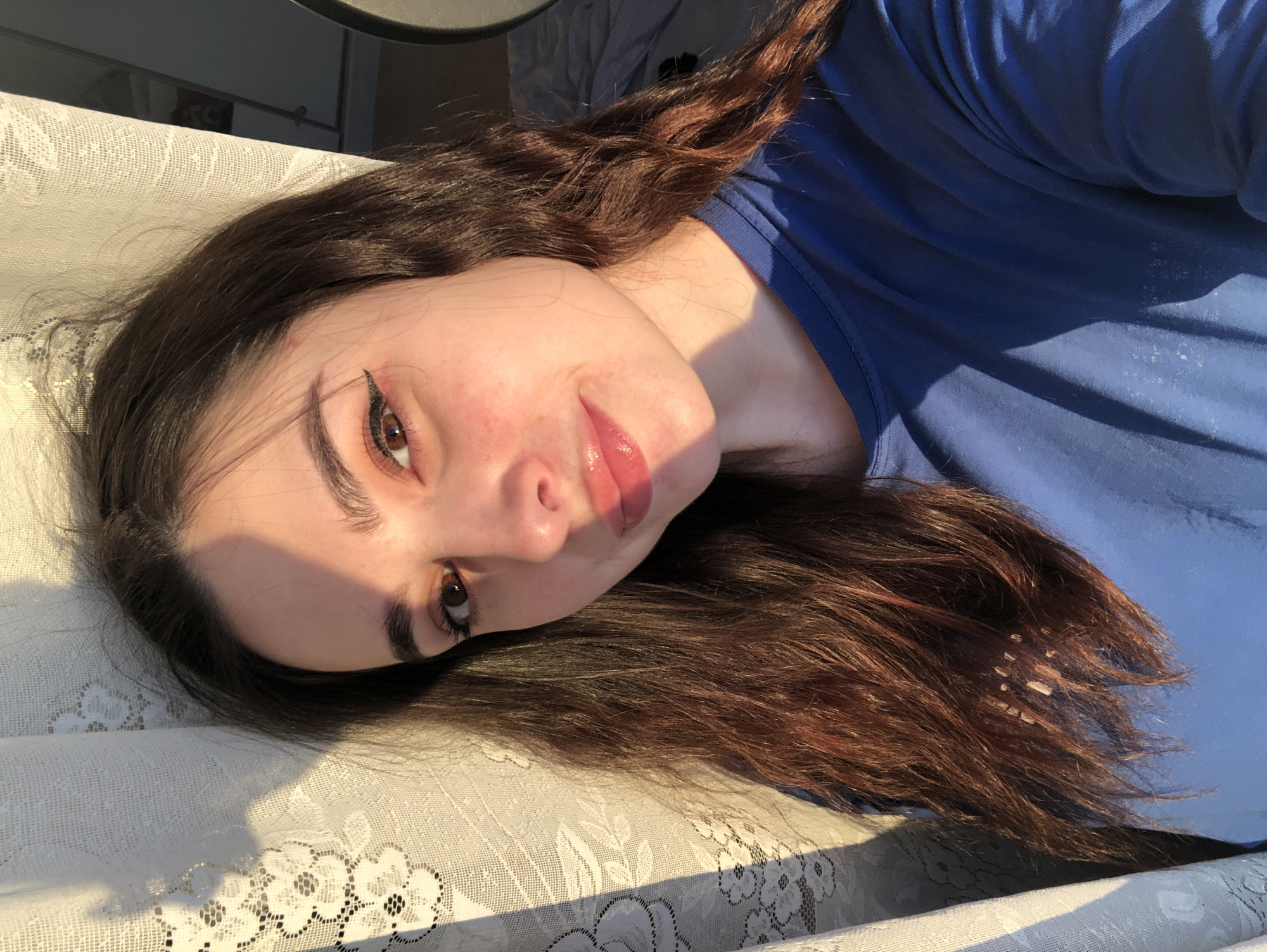 A young woman with long, wavy, auburn-brown hair and fair skin is lying on a white lace tablecloth. She is wearing a blue shirt. Her head is tilted towards the camera. She has brown eyes and winged eyeliner. Her lips appear to be coated in gloss. The scene is illuminated by sunlight, creating harsh shadows across the tablecloth and her clothing. The location is not discernible from the image, and the claim about Noviy, Russia cannot be verified.
