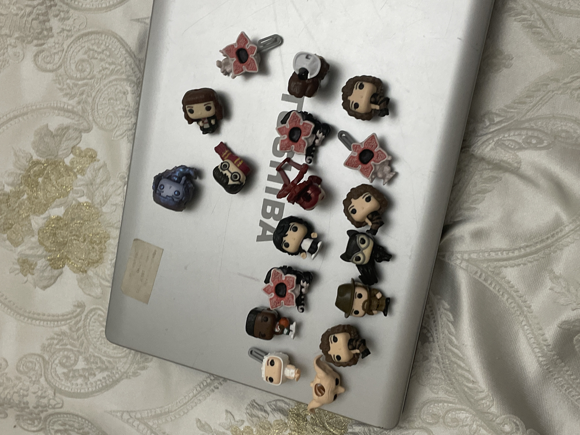The image captures a collection of small, stylized miniature figurines, likely Funko Pop! keychains or mini figures, arranged across the surface of a silver laptop. The laptop is positioned on a light-colored, patterned fabric, possibly a bedspread or tablecloth, which features an intricate floral and paisley design with subtle gold accents.
The figurines are the primary subjects, numbering around 18. Many of them appear to be characters from the "Stranger Things" series, including several Demogorgons with their distinctive open, petal-like heads, figures resembling Eleven (with short dark hair), Chief Hopper (distinguishable by his hat and beard), and Dustin. Other identifiable characters include one resembling Harry Potter (with glasses and a scarf), a blue-grey creature, a figure with owl-like characteristics, a character holding a chainsaw, and a pale, elongated monster.
The laptop itself is silver, showing some signs of use like smudges. A faded rectangular sticker is visible on its lower-left side. Partially visible text, "BA," can be seen on the laptop's surface, potentially part of a "TOSHIBA" brand logo or other text. The arrangement of the figurines suggests a static display rather than an active event.
The setting is indoors, implied by the patterned fabric, likely a domestic environment. No specific time of day, weather, or emotional state is discernible from the image.