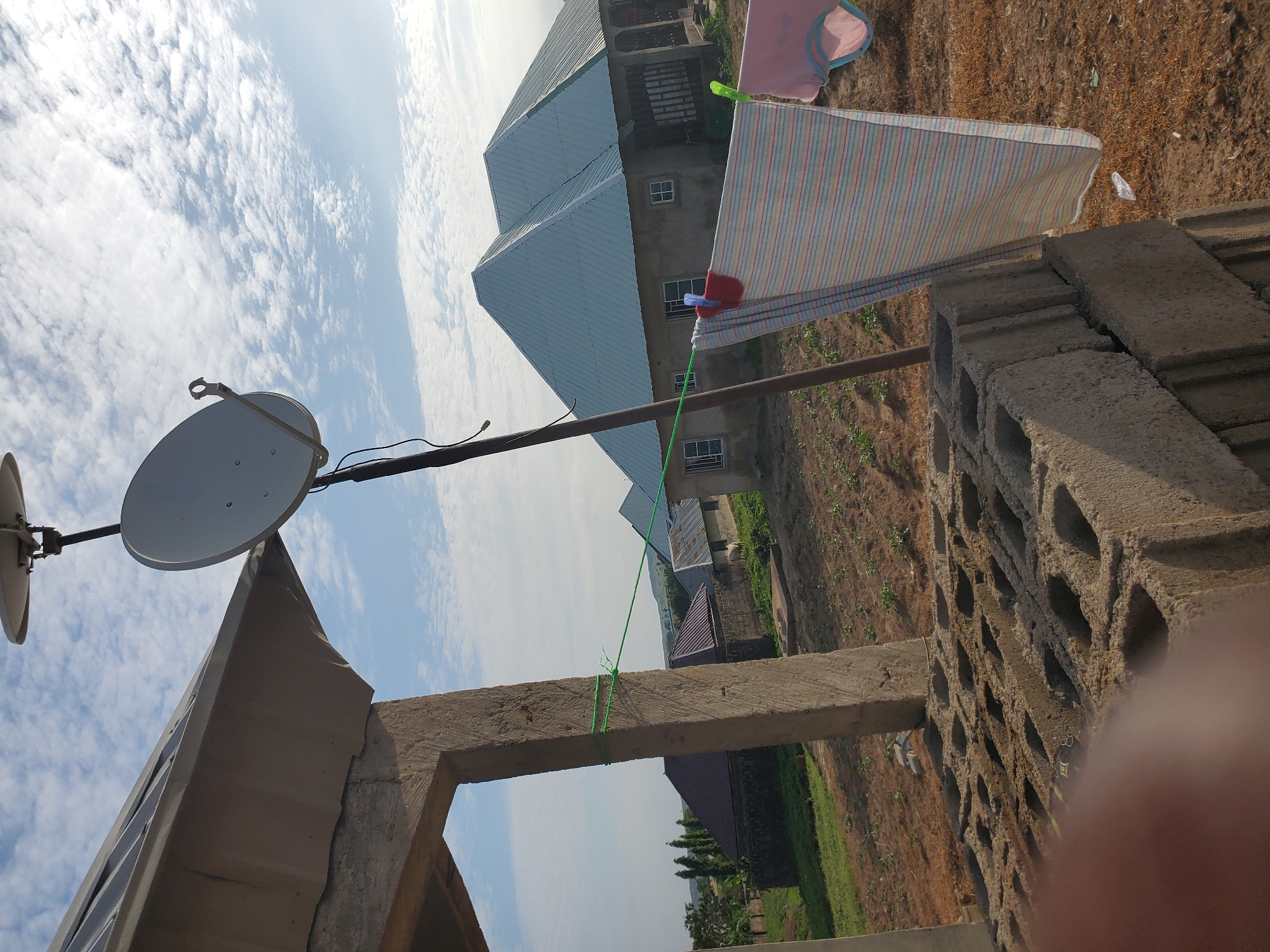 In the city of Winters, United States of America, an outdoor scene unfolds under a bright, partly cloudy sky. In the immediate foreground on the left, a stack of hollow concrete blocks (cinder blocks), approximately seven courses high, rests on dry, reddish-brown earth. A blurred, dark, fleshy object, likely a human finger, partially obstructs the lower-left corner of the frame.
A robust, unfinished concrete pillar, roughly square in cross-section and appearing to be part of a larger structure, stands vertically on the left side, extending from the bottom to the mid-top. A green nylon rope is tied securely around this pillar at two points and stretches horizontally to the right, functioning as a clothesline. Two items of laundry are suspended from this rope: a multi-striped, light-colored fabric (possibly a towel or sheet) on the left, fastened with a red clothespin, and a pink garment (appearing to be underwear or a small clothing item) on the right, secured with a light green clothespin.
Further into the midground, to the right of the concrete pillar, a tall, dark pole supports a gray parabolic satellite dish, with a smaller, secondary antenna-like component mounted above it. In the central midground, a large, light-colored house with a multi-pitched roof covered in light green/teal corrugated metal is prominent. The house features several small, rectangular windows, each divided into four panes, and its exterior walls appear to be unpainted concrete or stucco. The ground surrounding this house is dry, showing sparse vegetation and patches of bare earth.
To the left of the main house, another building with a darker, possibly maroon-colored, corrugated metal roof is visible, partially obscured by a cluster of tall, slender green trees. Adjacent to this, a smaller structure with a plain gray corrugated roof is present. Near this smaller building, on the dry ground, a light-colored animal, consistent with a goat or a small bovine, stands. The distant background reveals a hazy horizon with low-lying hills or mountains under a sky dotted with scattered, wispy white clouds.