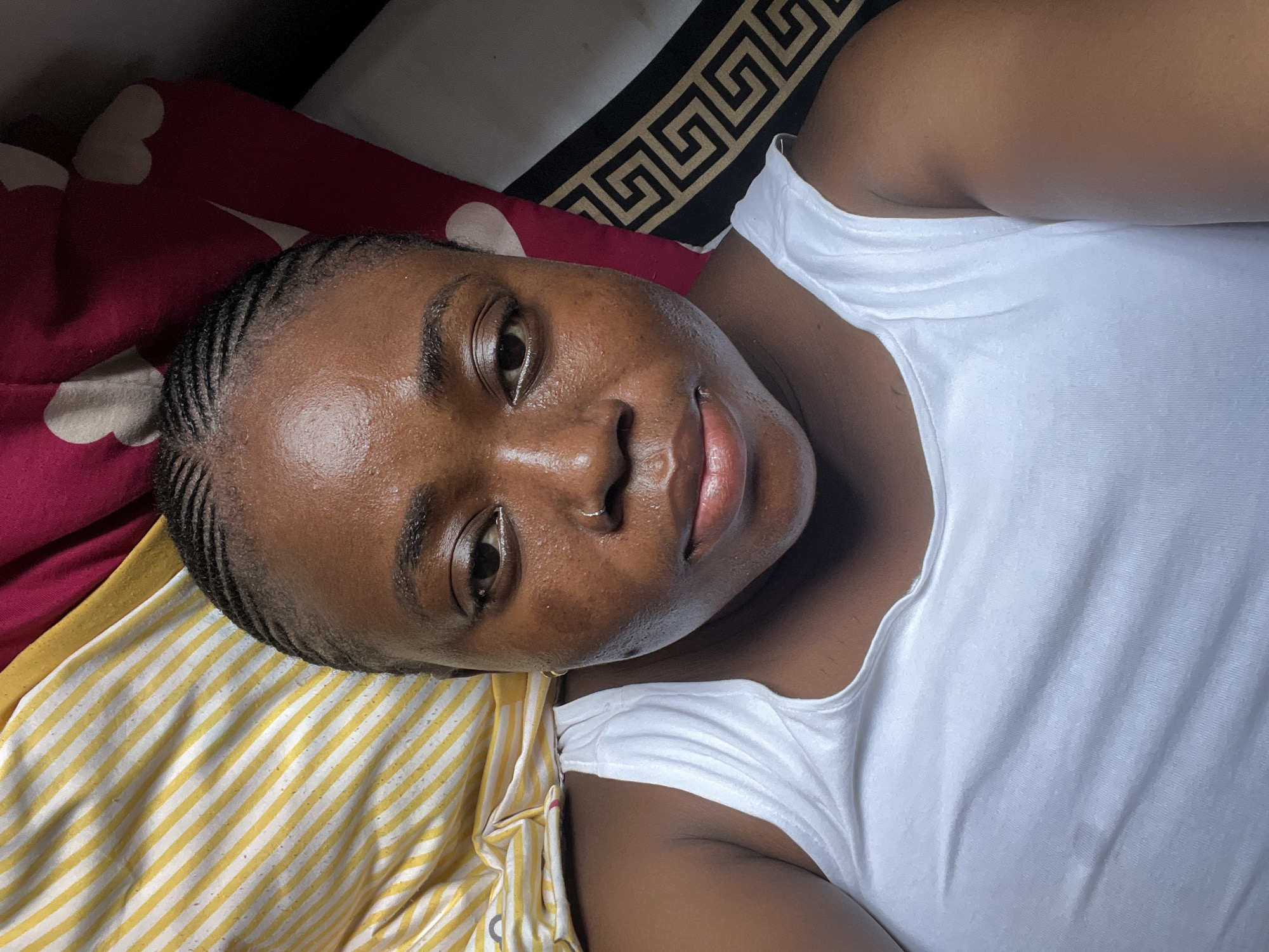This is a close-up, high-angle selfie of a Black woman lying down. She is wearing a white tank top and her dark hair is styled in small cornrows. The background consists of bedding with a yellow and white striped pattern and a pink fabric with white heart shapes, suggesting an indoor setting. Her face is the primary focus, with visible skin texture and subtle makeup. The image appears to have been taken in a bedroom environment, consistent with a residential area in Abuja, Nigeria.