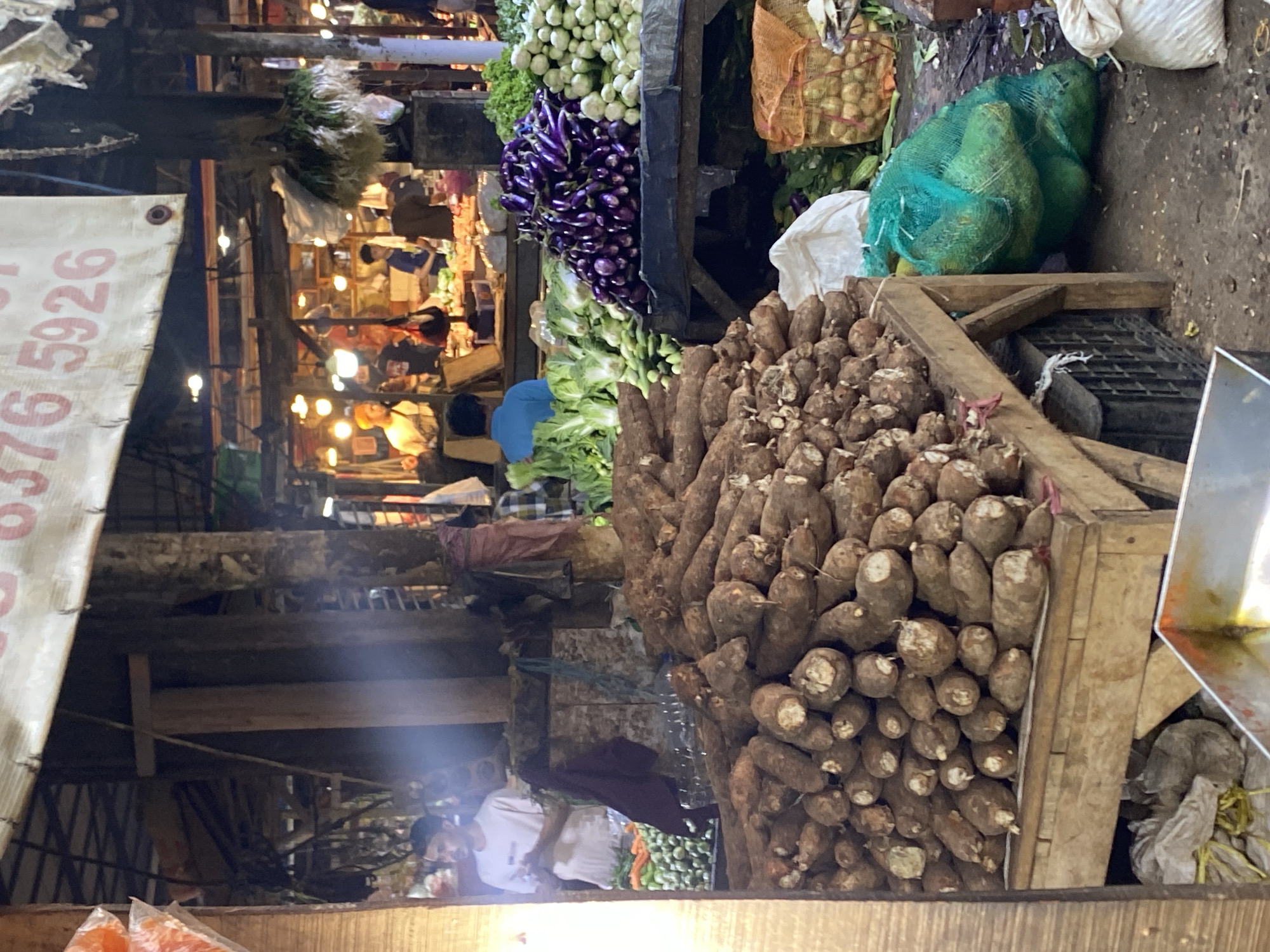 The image depicts a bustling outdoor market, likely in Cikupa, Indonesia, with a focus on fresh produce. In the foreground, a large pile of yams or cassava roots is displayed on a wooden table. To the right, a green mesh bag is filled with what appear to be large gourds or melons. Further back, various vegetables, including eggplants and leafy greens, are arranged on tables. Several individuals are visible amidst the market stalls, some attending to the produce and others browsing. The market appears to be covered by a corrugated metal roof, and artificial lighting illuminates the scene. A banner with red text and numbers, possibly indicating prices or a store name, is suspended overhead. The overall environment suggests a lively and active marketplace.