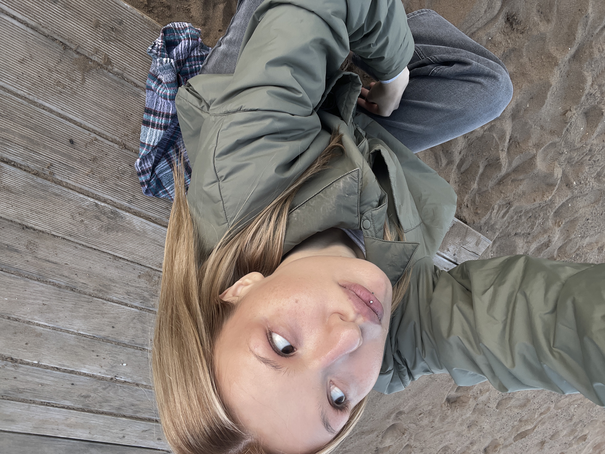 A young woman sits cross-legged on wooden decking, partially covered by sand, at a location with views of sand. She wears gray jeans and an olive-green jacket. A plaid blanket rests beside her. Her hair is blonde, and she has a lip piercing on the left side of her lower lip. She is looking upwards towards the camera. The environment suggests a beach or coastal area. The exact location in Chaikovsky, Russia, cannot be determined solely from the visual information provided.