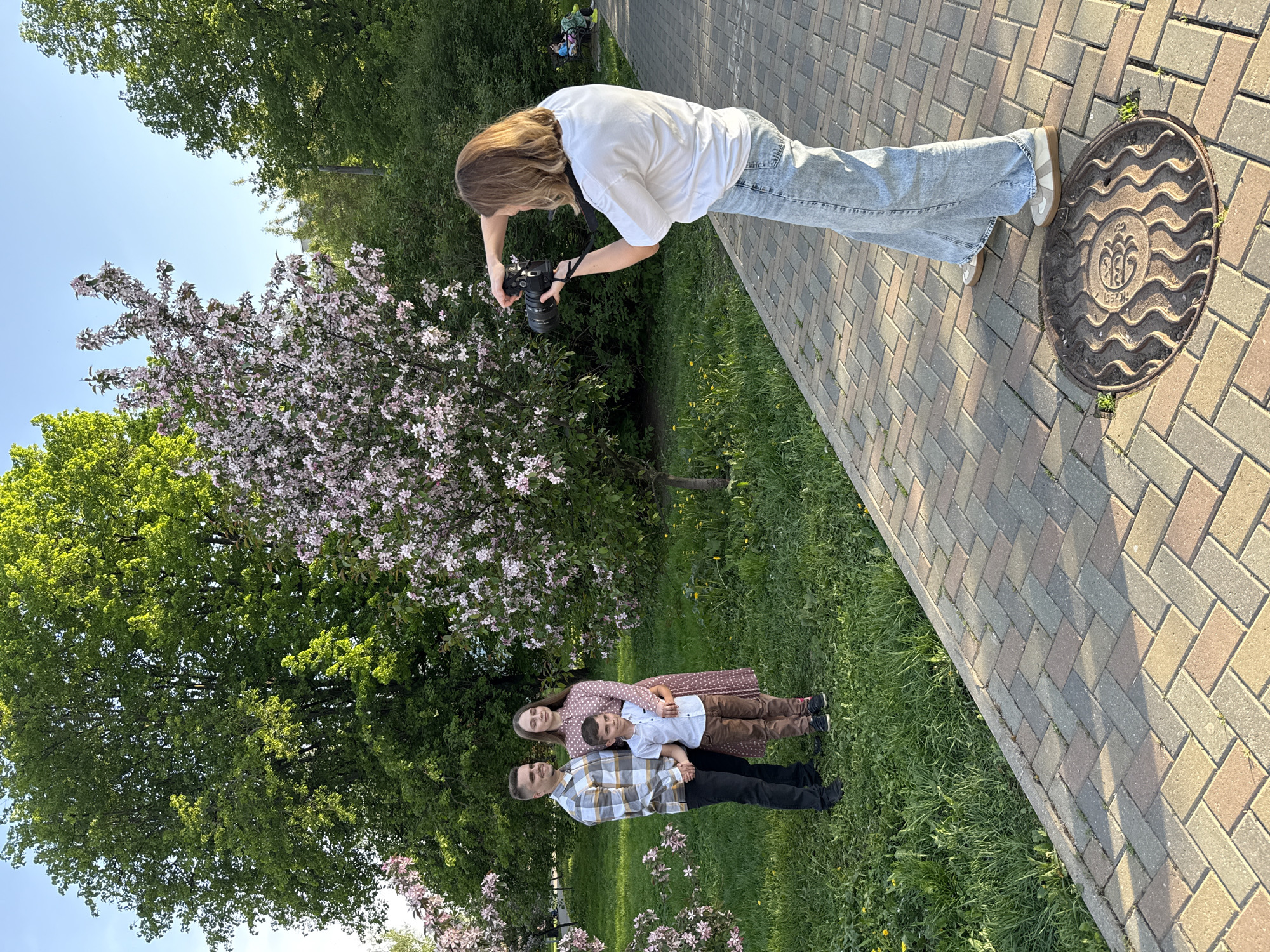 A woman with long brown hair stands on a brick-paved path in Rusanovo, Russia, holding a DSLR camera and appearing to take a photograph. She wears a white t-shirt, light blue jeans, and beige and white shoes. To her left, on a patch of green grass beneath blooming trees, a family of three poses. The family consists of a man in a plaid shirt and dark pants, a woman in a polka dot dress, and a boy in a white shirt and brown pants. The ground is grassy and contains scattered small yellow flowers. To the right of the woman with the camera is a manhole cover. The sky above is partly visible and blue.