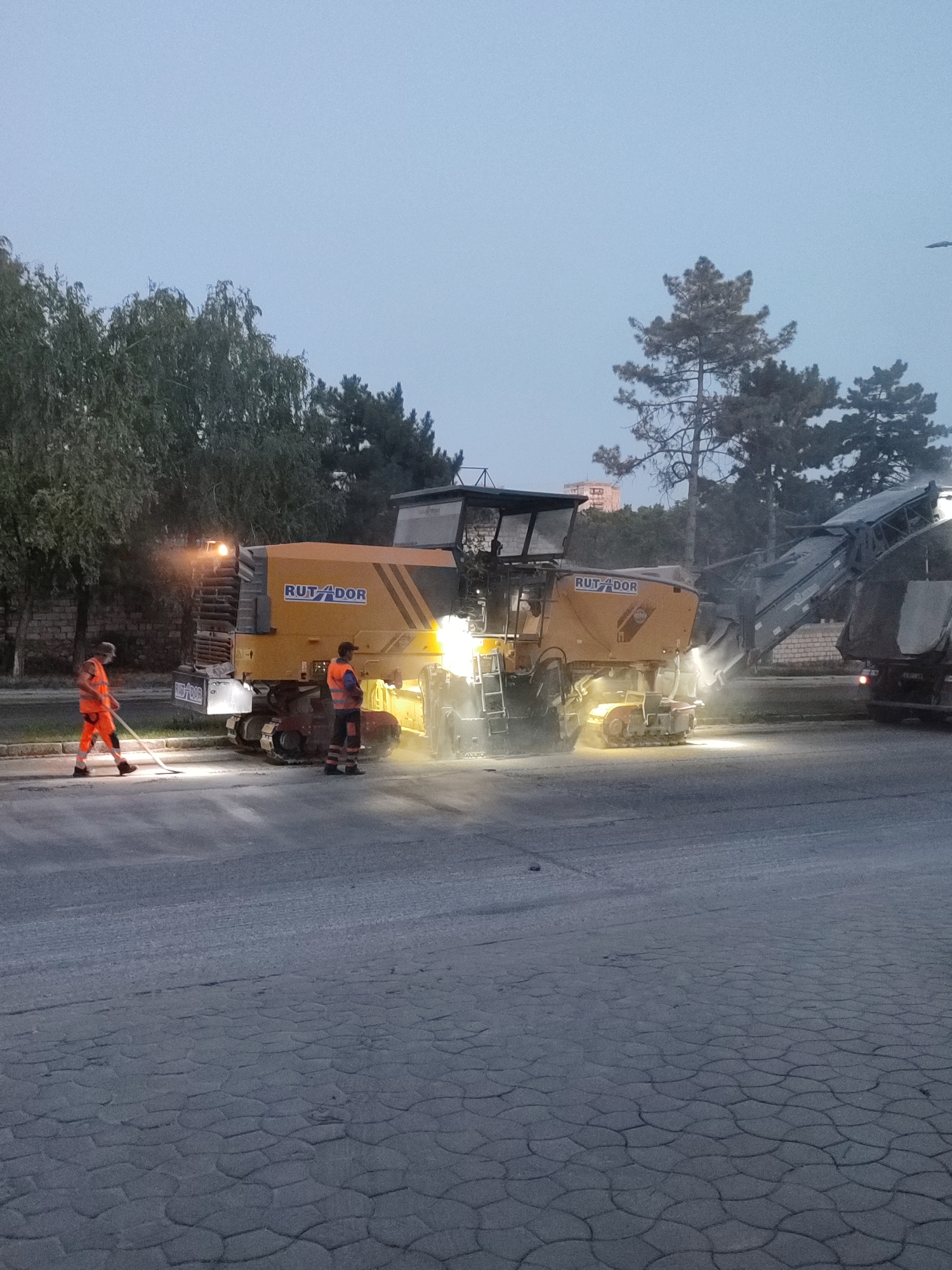 An outdoor scene in Chișinău, Moldova, depicts roadwork in progress, possibly in a park or pedestrian area, given the presence of what appears to be patterned paving stones in the foreground and mature trees lining the background. The lighting suggests either early morning or late evening.
The central focus is a large, orange road milling machine, branded "RUT-DOR." The machine is actively engaged in milling the asphalt surface, indicated by dust and debris being expelled. Two construction workers, dressed in high-visibility orange vests and trousers, are present. One worker is positioned to the left of the milling machine, holding a broom and appearing to sweep the debris. The other worker stands to the right of the machine, facing it. The milling machine is illuminated by its own lights, casting a bright glow on the surrounding area.
In the background, more trees are visible, with a building in the distance. The sky is a muted, overcast grey. A large truck is partially visible to the far right, likely involved in the roadwork operation, possibly for material transport or debris removal. The overall impression is one of industrial activity taking place in a somewhat urbanized natural setting.