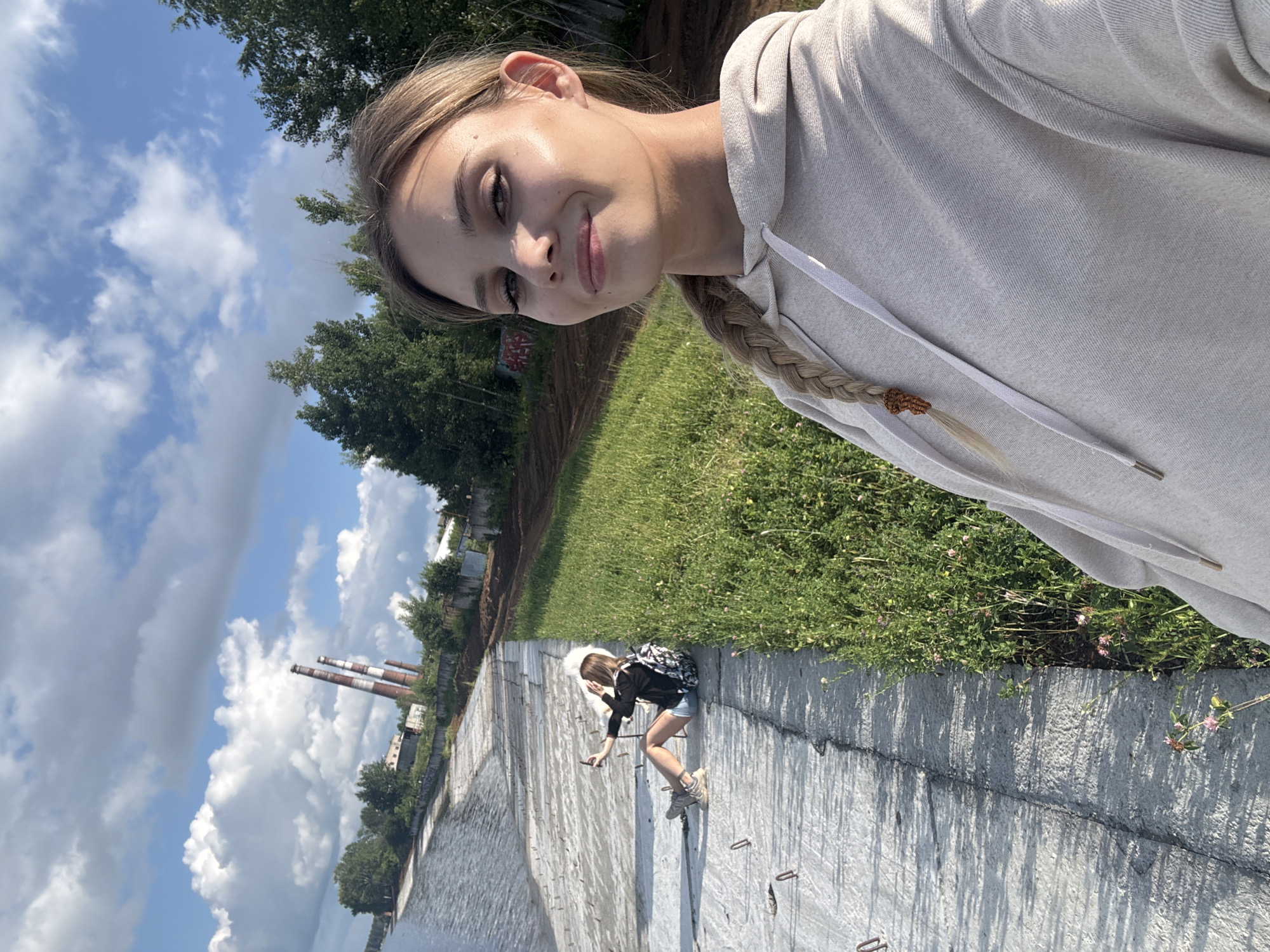 The image, taken in Chaikovsky, Russia, depicts two young women by a body of water, possibly a canal or river, with a concrete embankment. The woman in the foreground is taking a selfie, smiling and looking towards the camera. She has blonde hair braided in a plait and is wearing a light-colored hoodie. In the background, a second woman is seated on the concrete embankment, holding a phone and appears to be taking a photo or video. She has blonde hair and is wearing a dark jacket and shorts, with a backpack next to her. The environment features a grassy slope leading up from the embankment, with trees and industrial smokestacks visible in the distance under a partly cloudy sky.