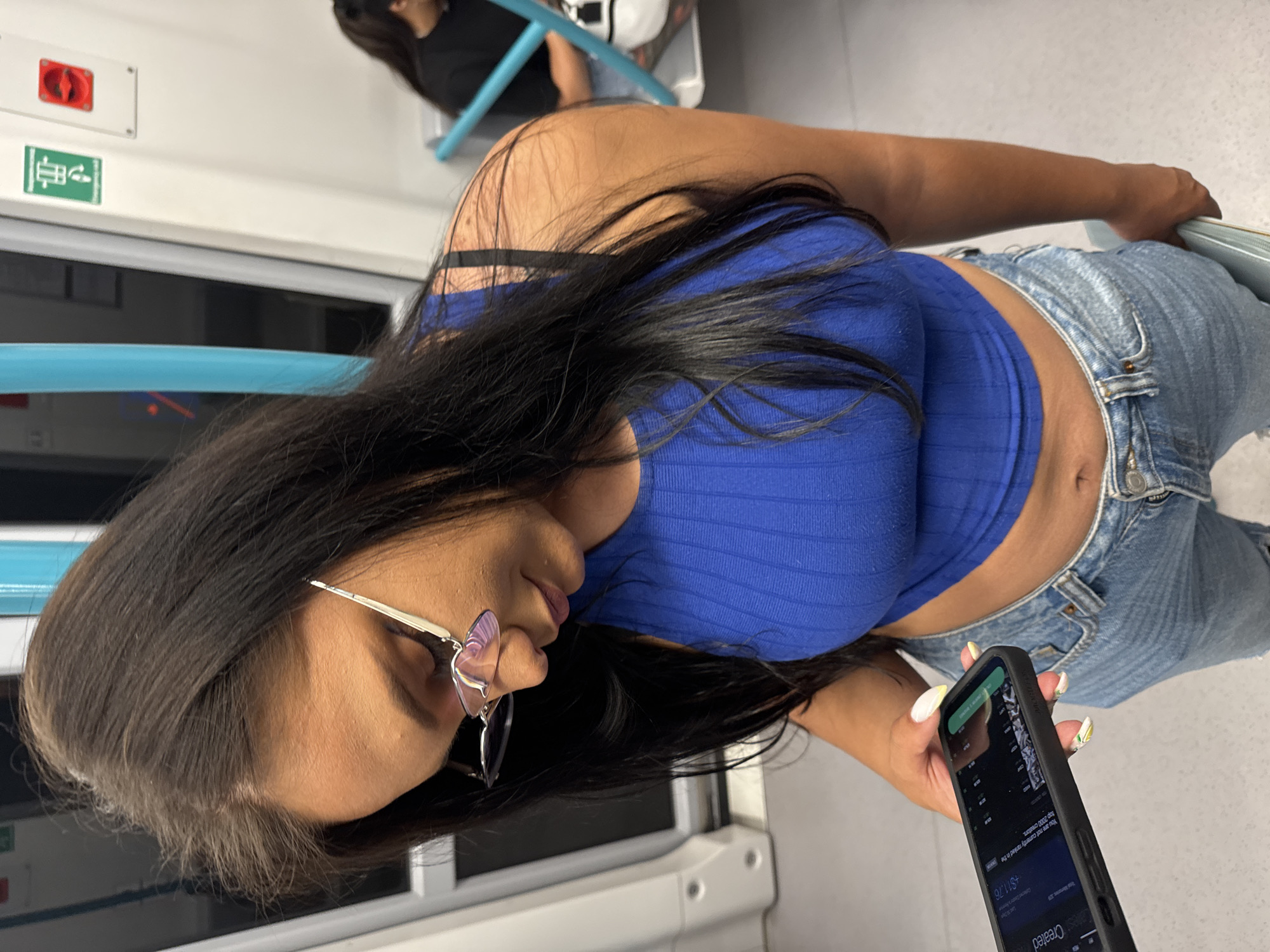 A woman with long, dark hair and glasses, wearing a blue sleeveless top and denim jeans, is shown holding a smartphone in her hand. She appears to be on public transportation, possibly a train, given the presence of seats, poles, and windows typical of such an environment. The woman is looking down at her phone, which displays a screen with text and graphics. The background shows a blurred interior of the vehicle, with another passenger visible in the distance. The scene is indoors, in what appears to be a dimly lit setting. There is no visible indication of the city of Sofia, Bulgaria.