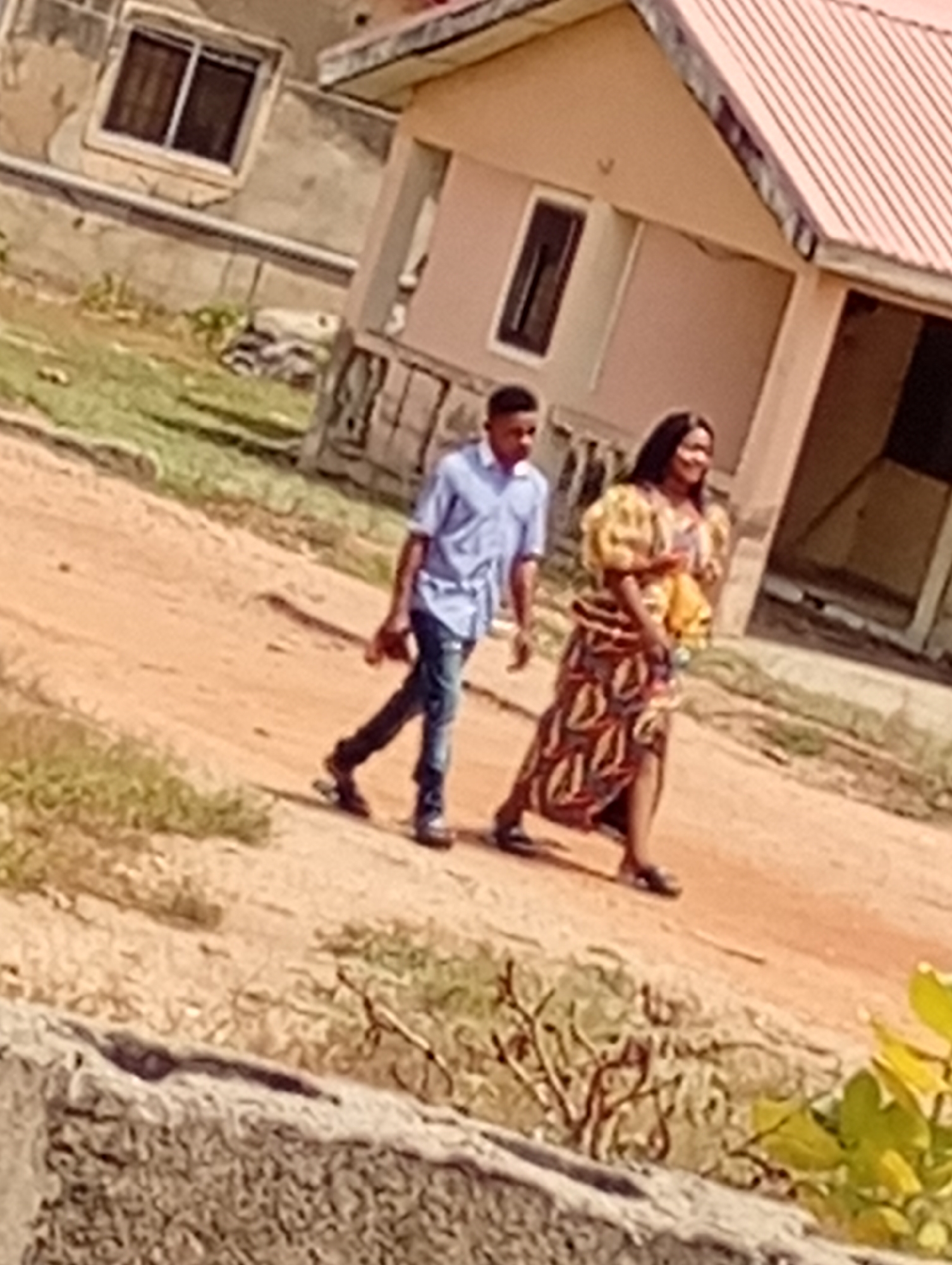 A man and a woman are walking on a dirt path outside a residential building in an area identified as Unknown, Unknown. The man is wearing a blue button-down shirt and jeans. The woman is wearing a patterned dress with puffy sleeves. The building has a window and a red roof. There is vegetation along the path.