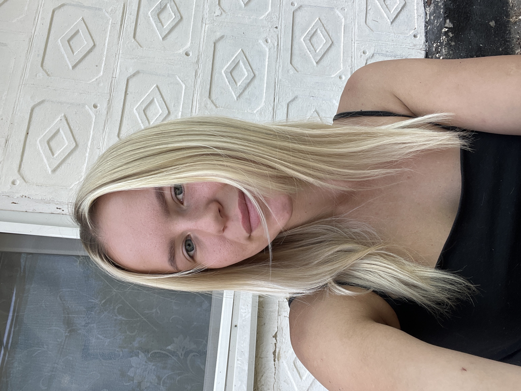A blonde woman with blue eyes is pictured in a medium close-up selfie. She is wearing a black tank top and her hair is styled straight. She is positioned in front of a white brick wall with a patterned texture, and to her left, there is a window with a screen. The lighting suggests an outdoor setting during the daytime.
