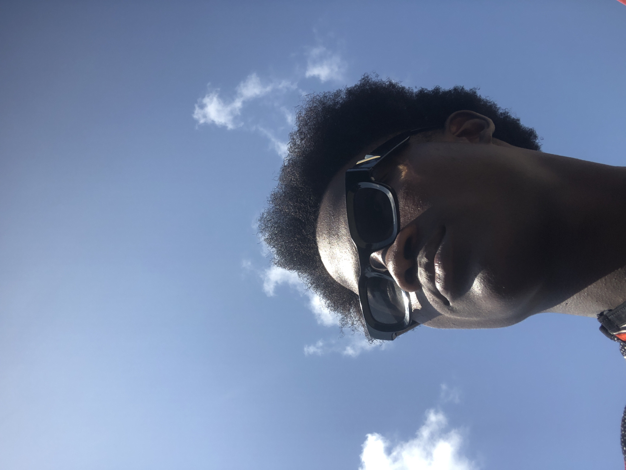 This media file features a striking, low-angle portrait of a person looking upwards against a vibrant blue sky dotted with scattered white clouds. The subject, seen primarily from the chin to just above the head, has dark-toned skin and short, tightly curled dark hair. They are wearing prominent dark, rectangular sunglasses that conceal their eyes and much of the upper face. The strong overhead or backlighting creates a dramatic effect, rendering the face largely in shadow, almost in silhouette, while highlighting contours on the forehead, bridge of the nose, and lips. The close-up perspective emphasizes the textures of the hair and skin. A glimpse of an ear is visible on the left side of the frame, near the hairline. On the lower right, a small fragment of a red, white, and blue patterned strap or collar is visible. The bright, clear sky suggests it is a sunny day with fair weather. No text is discernible in the image. This outdoor scene is set in Ado, Nigeria.