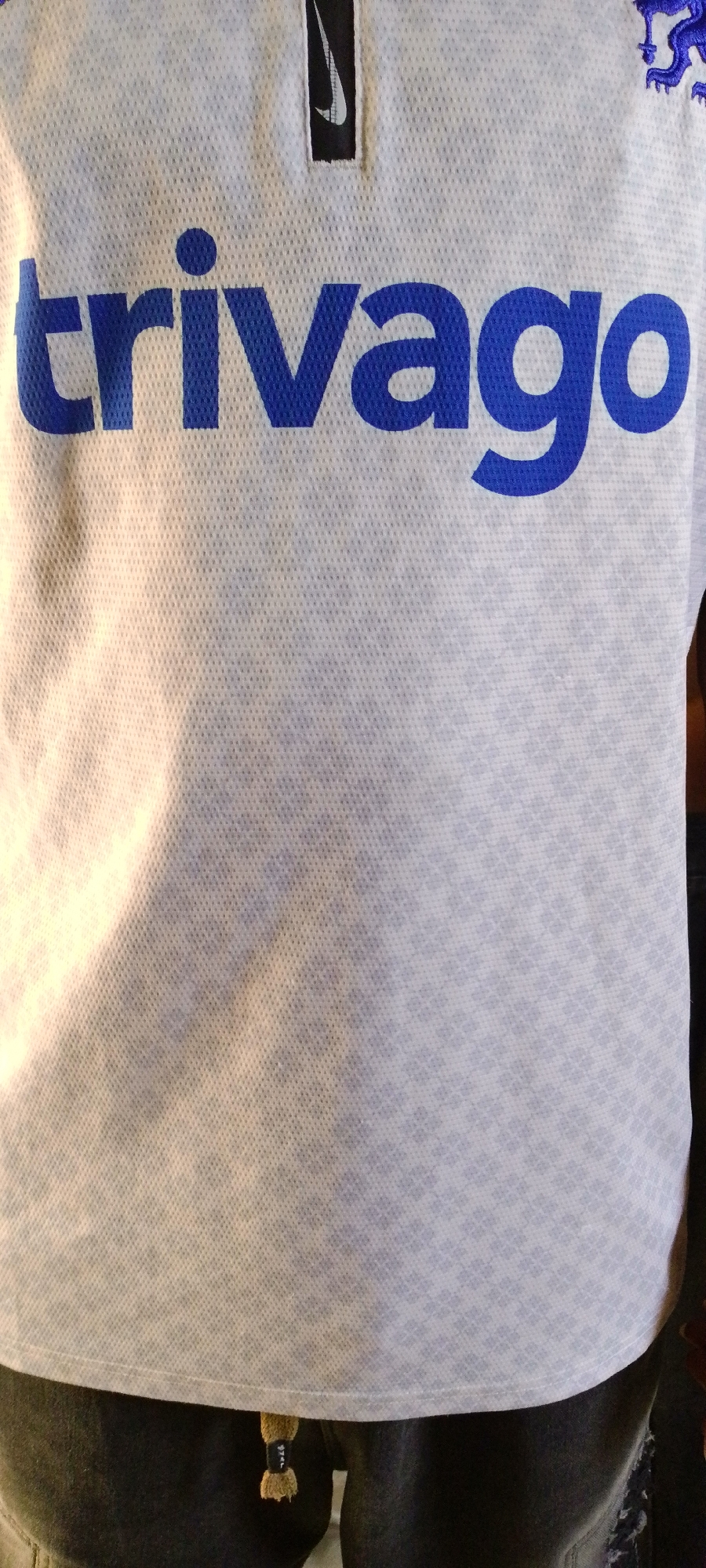 The image shows a close-up of a white t-shirt with a blue logo that reads "trivago". The t-shirt also features a black Nike logo near the collar. The fabric of the t-shirt has a subtle checkered pattern. Below the t-shirt, dark pants are visible. A small brown tag with black lettering is attached to the pants. The lighting suggests an indoor setting. There is no indication of the city of Jalingo, Nigeria, or any specific urban environment.
