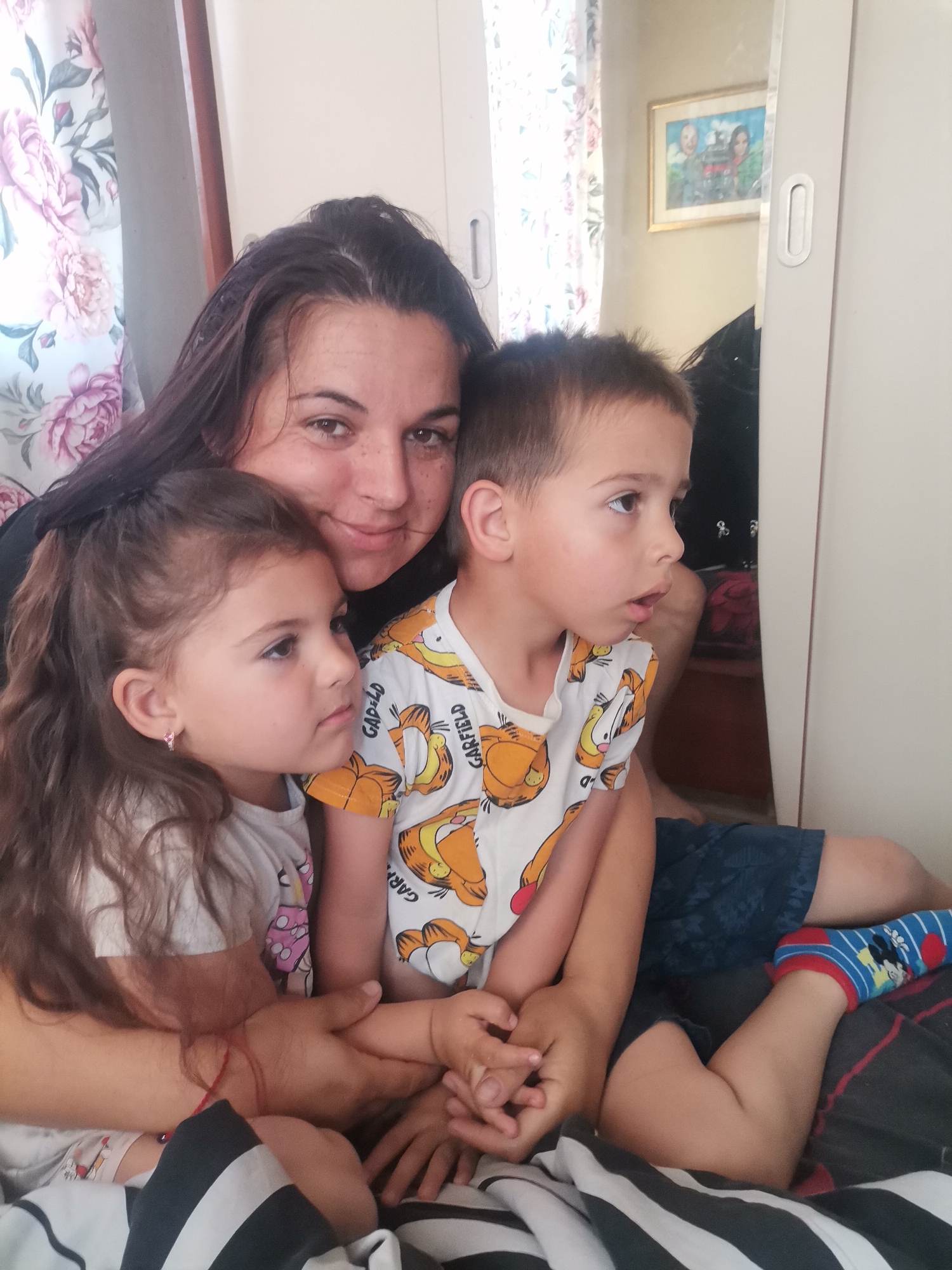 In an indoor setting, likely a bedroom or living area, a woman is seen with two young children, a girl and a boy. The woman, with dark hair and a warm smile, looks directly towards the camera, her arm around the young girl. The girl, with long dark brown hair styled in a half-ponytail and wearing a white top and small earrings, looks intently to her right, away from the camera. Beside her, a young boy with short light brown hair, dressed in a white t-shirt adorned with "GARFIELD" text and blue shorts, also looks in the same direction with a slightly open mouth, appearing focused. The woman wears a dark top and has a red string bracelet on her left wrist. The boy has one foot visible, wearing a blue sock with a Mickey Mouse design.
The background features floral curtains with pink and purple patterns on the left. To the right, a white wardrobe or cabinet with a reflective surface is visible. Reflected in the mirror is a framed picture, depicting what appears to be a portrait of three individuals. The family is seated on a bed covered with black and white striped bedding. The scene suggests a relaxed daytime moment, possibly a family relaxing or watching something together. The emotions conveyed are warmth and engagement, with the woman appearing affectionate and the children engrossed in something outside the frame. The specific location remains unknown.