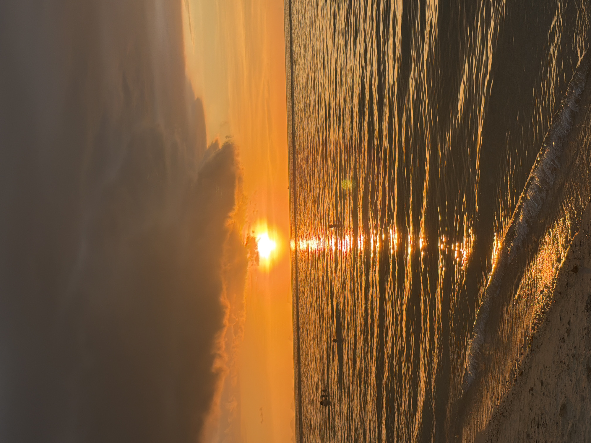 The image depicts a sunset over the ocean in Baan Tai, Thailand. The sun, a bright orb, is partially obscured by dark clouds, casting a warm, golden glow across the water and sky. The ocean surface is rippled, reflecting the sunlight in a shimmering path towards the viewer. Several individuals are visible in the water, appearing to be swimming or wading. The foreground shows a sandy beach with gentle waves lapping at the shore. The overall atmosphere is serene and tranquil, capturing the beauty of a tropical sunset.