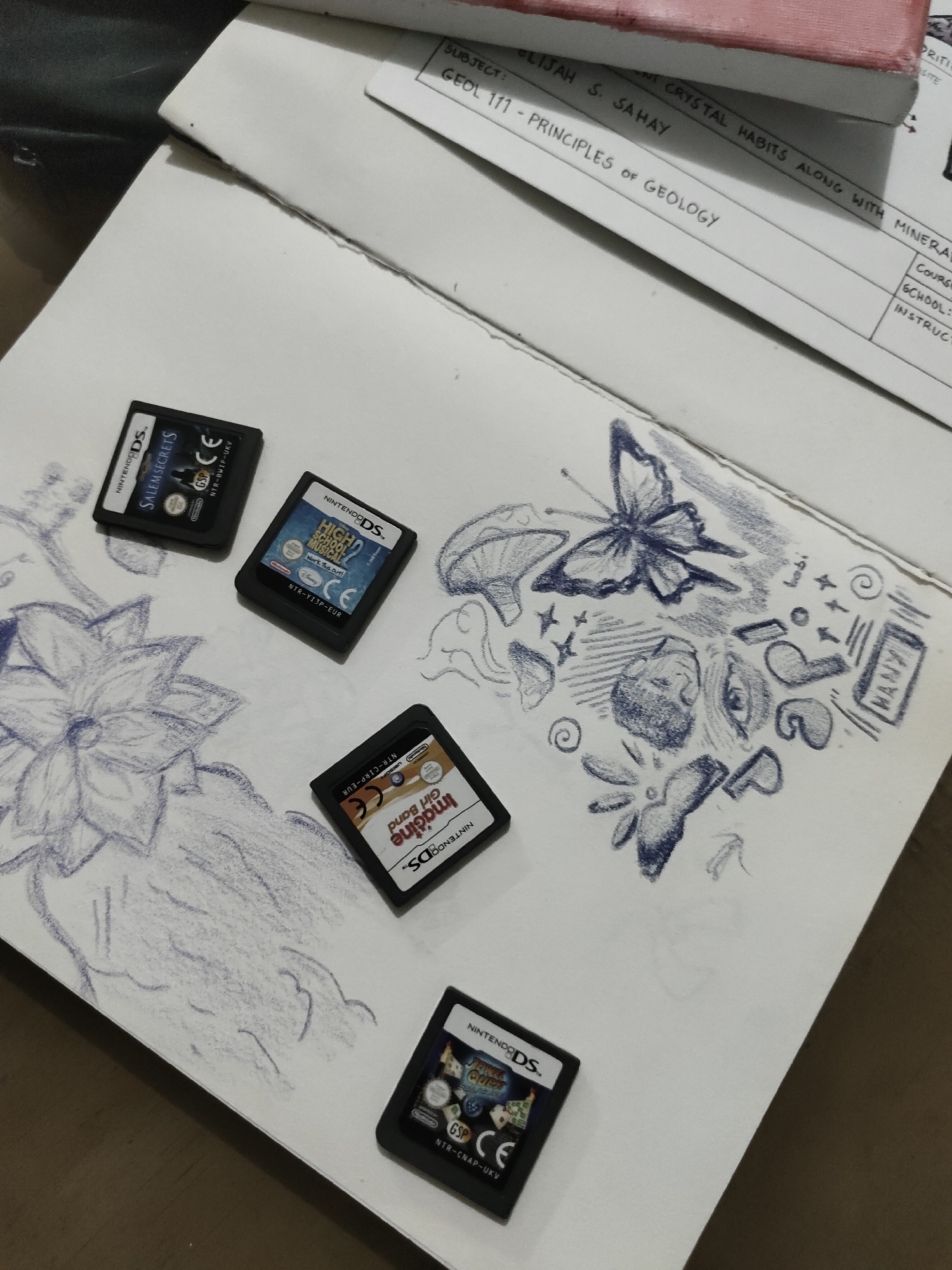 The image displays an overhead shot of four Nintendo DS game cartridges, an open notebook with blue pencil sketches, and a closed academic notebook, all resting on a light-colored surface, likely a desk or table, in Davao City, Philippines.
In the foreground, four black Nintendo DS cartridges are visible. From left to right and top to bottom, the games are identified as "Salem Secrets," "High School Musical 2: Work This Out!", "Imagine Girl Band" (oriented upside down), and "Jewel Quest Expeditions."
Beneath and around the game cartridges, an open white notebook reveals blue pencil drawings. On the left side of the notebook, a detailed sketch of a large, multi-petaled flower dominates the page. On the right side, a collection of smaller, whimsical doodles is present, including a butterfly, a mushroom, an eye, various abstract shapes, swirls, stars, and the discernible word "MANY."
Positioned above the open notebook and partially obscured is a closed academic notebook or book with a reddish spine. A white paper, likely a syllabus or assignment sheet, is placed on top of it. Text on this paper indicates "SUBJECT: GEOL 1/1 - PRINCIPLES OF GEOLOGY" and "ELIJAH S. SAHAY." Other visible fragments include "CRYSTAL HABITS ALONG WITH MINERAL..." and parts of labels like "COURSE," "SCHOOL," and "INSTRUCT."
The background is a dark, indistinct surface. No people are visible in the scene. The items suggest a personal study or leisure setting.