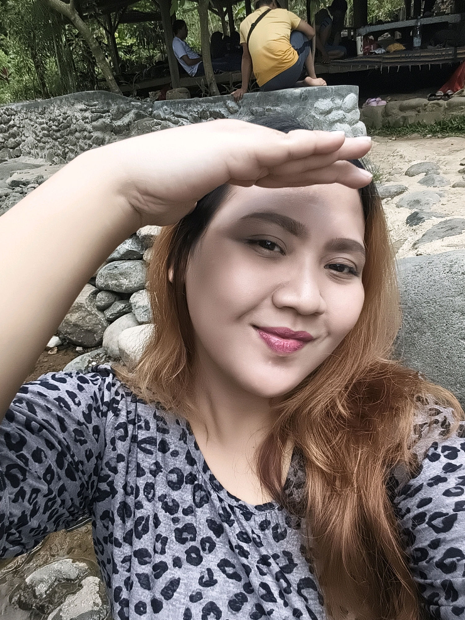 In a selfie taken outdoors, a woman with long, auburn hair and pink lipstick shields her eyes from the sun with her right hand, which is raised above her forehead. She is wearing a gray shirt with a black leopard print. The background appears to be a rocky, natural setting with some trees and a covered wooden structure in the distance. In the background, to the left, a stone wall is visible. Several people are seated under the wooden structure, some appearing to be resting. The ground is covered in stones and dirt.