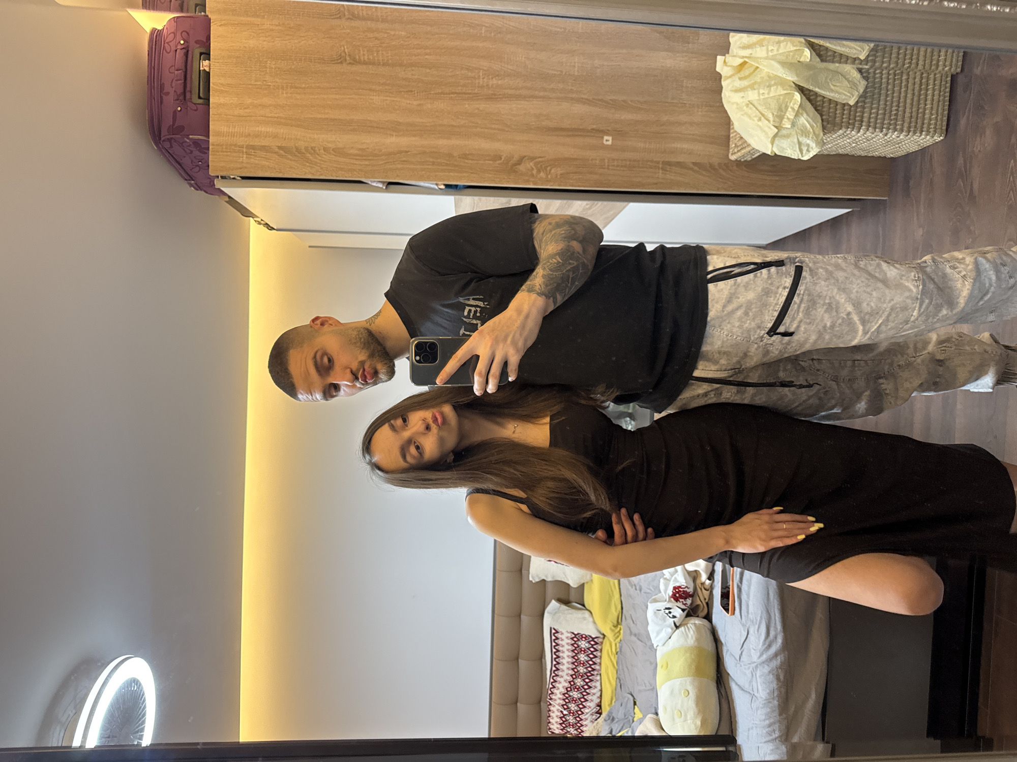 A man and a woman are posing for a selfie in front of a mirror. The man is bald with a beard and is wearing a black t-shirt and gray pants. He is holding a phone in his right hand. The woman has long brown hair and is wearing a black dress. She is placing her hand on her upper thigh. Behind them is a bed with pillows and blankets. A wardrobe can be seen behind them, with a suitcase sitting on top. A light ring is visible in the bottom left corner. The location appears to be a bedroom in Sofia, Bulgaria.