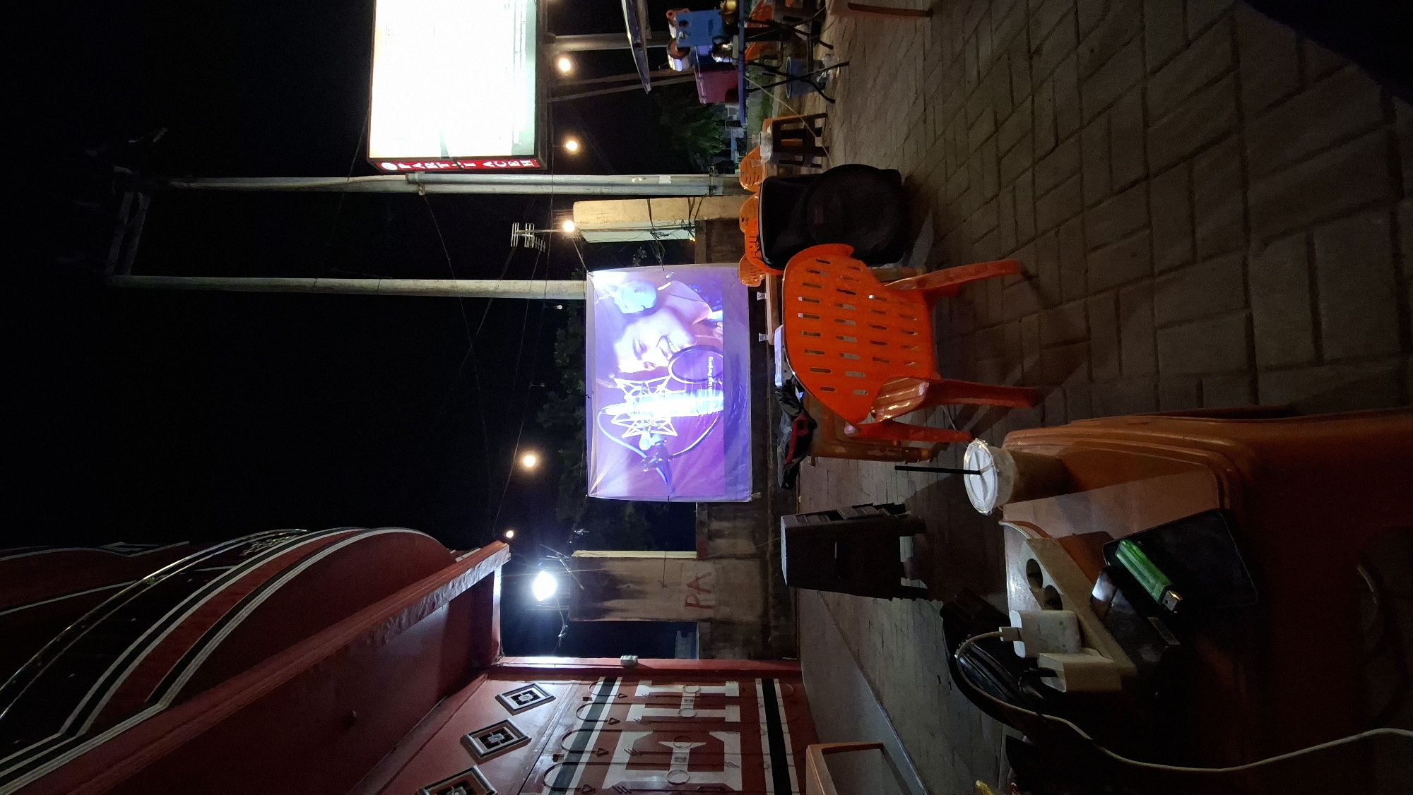 The image depicts an outdoor seating area at night, possibly a cafe or entertainment venue, located in Keudeteupinpunti, Indonesia. A large banner displaying a close-up of a smiling young man wearing headphones and singing into a microphone is projected onto a screen. Several orange plastic chairs are arranged in front of the screen, with some tables and stools nearby. On one of the tables in the foreground, a cup with a straw, a mobile phone, a lighter, and a power adapter are visible. To the left, part of a red and white building with intricate decorative patterns is visible. The ground is paved with dark brickwork. In the background, streetlights and poles indicate an urban setting. There are no visible people in the immediate foreground, but the setup suggests an area where people might gather to watch the projection.