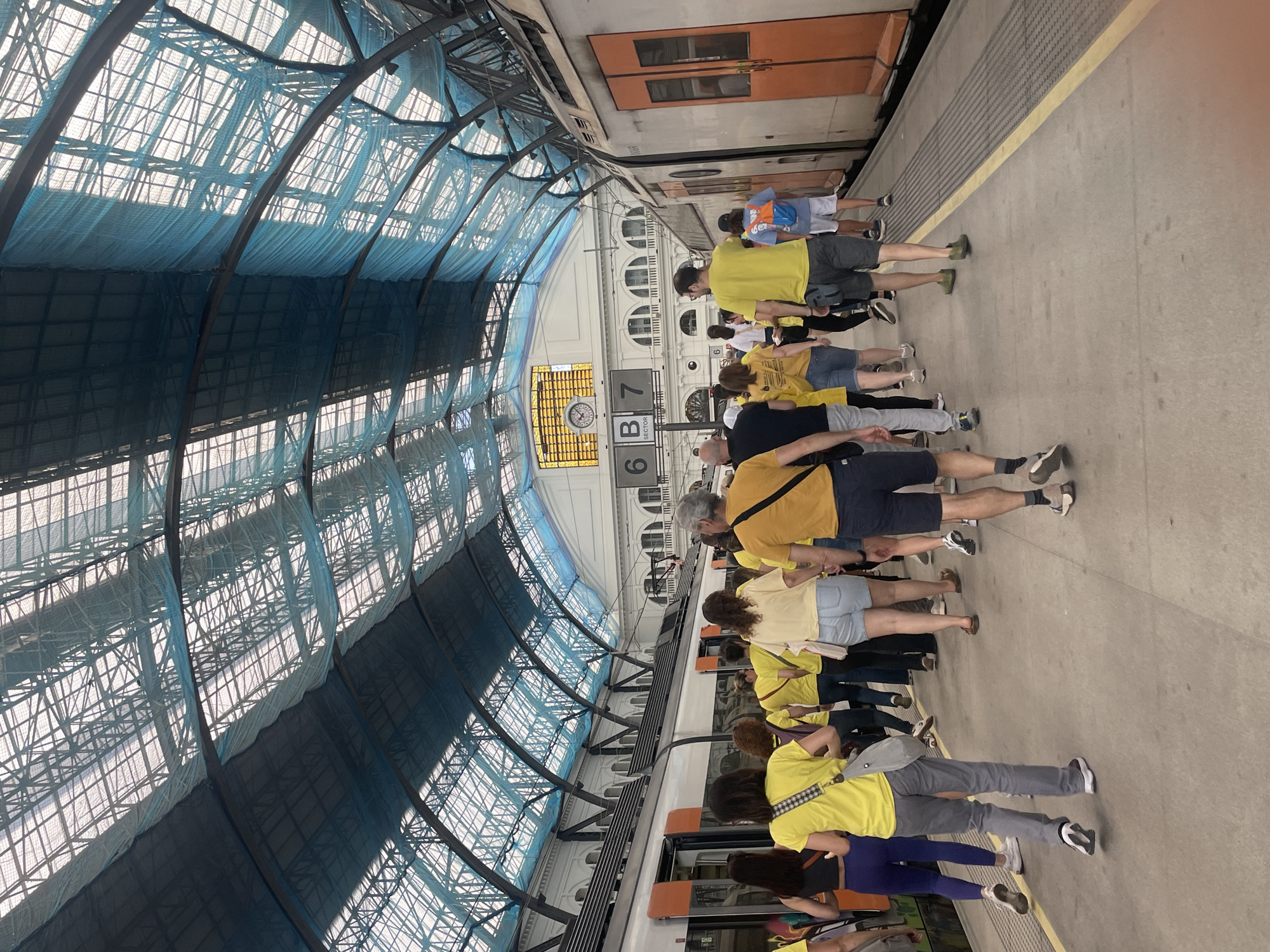 A photograph depicts a group of people standing on a train platform, presumably waiting to board a train. The group is predominantly wearing yellow shirts, and they are standing adjacent to a train. The architectural structure of the train station is visible, featuring a high ceiling with a complex network of metal beams and a blue-tinted netting. Signage indicates platform numbers, with '6' and '7' visible. The photograph is taken from an elevated perspective, and the angle of the image suggests it was not originally oriented in this way. The details present do not allow for definitive identification of the location as Barcelona, Spain, although the architectural style is consistent with European train stations.