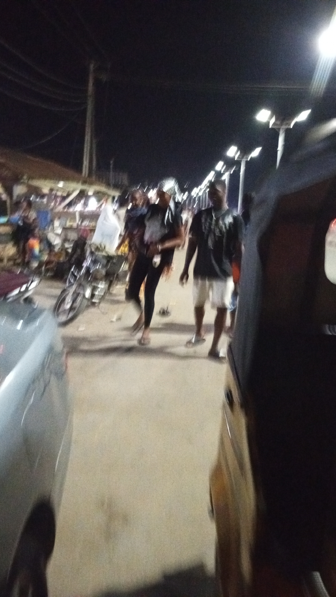 The image depicts a dimly lit street scene at night in Adankolo, Nigeria. Two individuals are the primary focus: one wearing dark pants and a light-colored top, and another wearing a dark shirt and light-colored shorts. Both appear to be walking along the street. A parked motorcycle is visible near a building or structure that appears to have stalls or vendors. Cars are partially visible on either side of the street. Streetlights illuminate the scene.