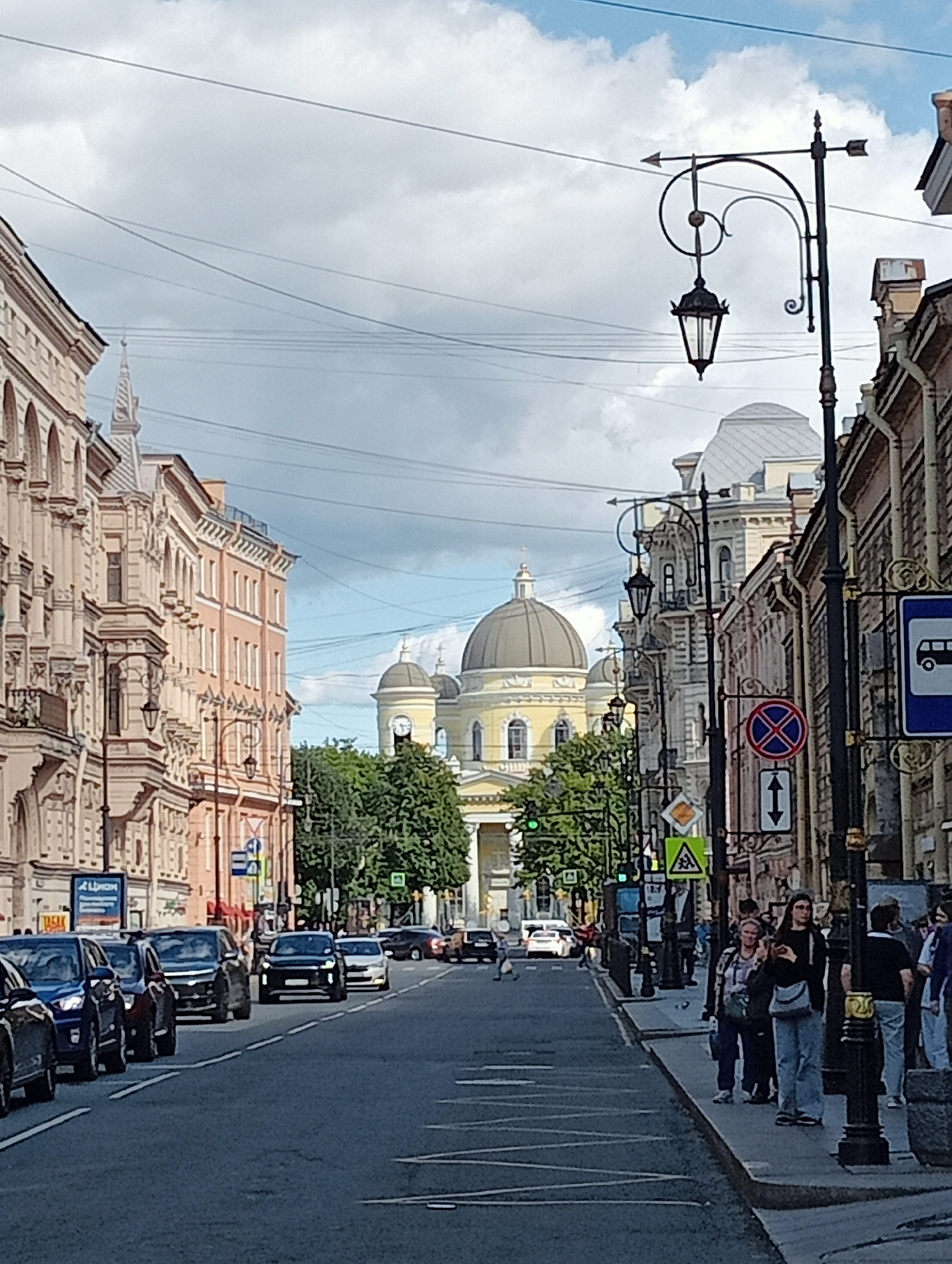 The image shows a view down Nevsky Prospekt in Saint Petersburg, Russia, with the Kazan Cathedral visible in the distance. The sky is partly cloudy, with a mix of blue sky and white clouds. A street lamp with a decorative design stands on the right side of the street, and there are several cars parked and driving on the road. Pedestrians are visible on the sidewalk. The buildings on either side of the street are of classical architecture, with ornate facades. Traffic signs are present, including a bus stop sign and a sign indicating no stopping. The overall impression is of a bustling city street scene.