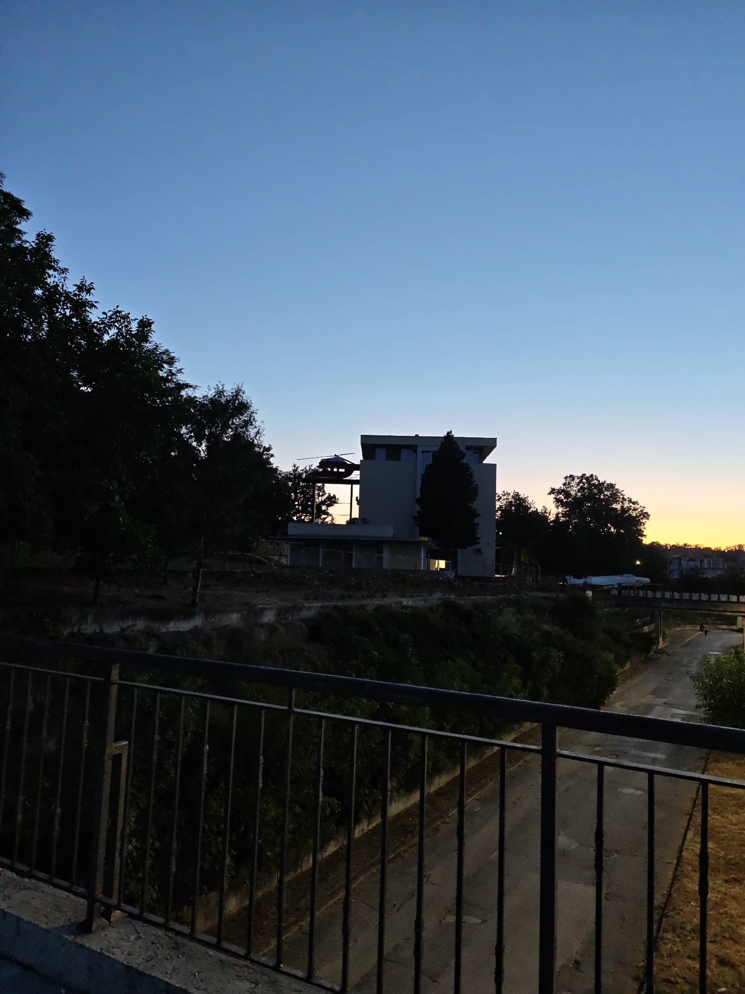 The image depicts a twilight scene in Pleven, Bulgaria, overlooking an area with a building and a road. In the foreground, a dark metal railing runs horizontally, suggesting the perspective is from a balcony or bridge. Beyond the railing, a steep, vegetated embankment is visible, with dense trees and shrubs.
Dominating the mid-ground is a modern, multi-story building with a distinctive angular roofline. The building is partially silhouetted against the fading light, with some windows reflecting the warm glow of sunset. A dark, helicopter-like object is perched on a platform or structure attached to the building, positioned at the upper left side of the building. A tall, dark evergreen tree stands to the right of the building.
To the right of the building, a road or pathway is visible, appearing to descend slightly. A simple concrete bridge crosses over this pathway further in the distance. The sky above is a gradient of deep blue at the top, transitioning to lighter blues and then a warm yellow-orange hue near the horizon, indicating the sun has recently set. Trees are visible on the horizon, further obscuring the landscape. The overall atmosphere is calm and serene, characteristic of early evening in an urban environment.