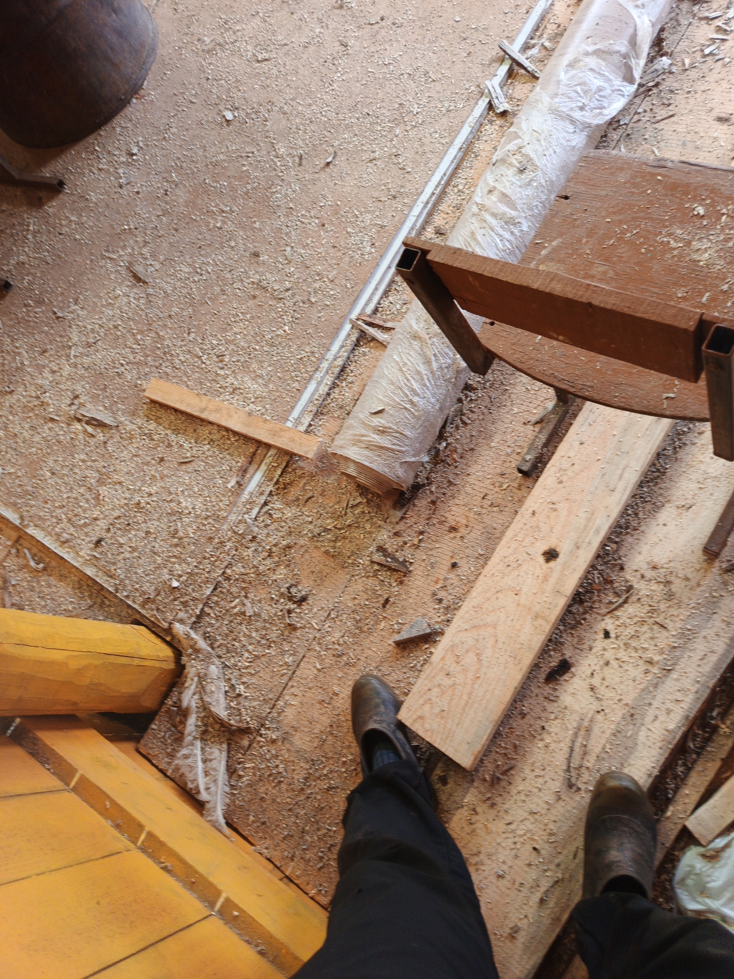 This image captures a floor covered in sawdust, likely in a workshop or construction area, located in the city of Islambaevo, Russia. A dark-colored, large cylindrical object, possibly a drum or container, is visible in the upper left corner. To the right of this object, a long, white, plastic-wrapped item is coiled on the floor. A rustic wooden table with metal legs is positioned in the mid-ground, with several wooden planks lying on and around it. In the foreground, a yellow-painted wooden structure, possibly part of a floor or wall, is visible on the left. The lower portion of the image shows a person's legs and feet clad in dark pants and boots, standing on the sawdust-covered floor. The overall environment appears to be utilitarian and reflects an active work area.