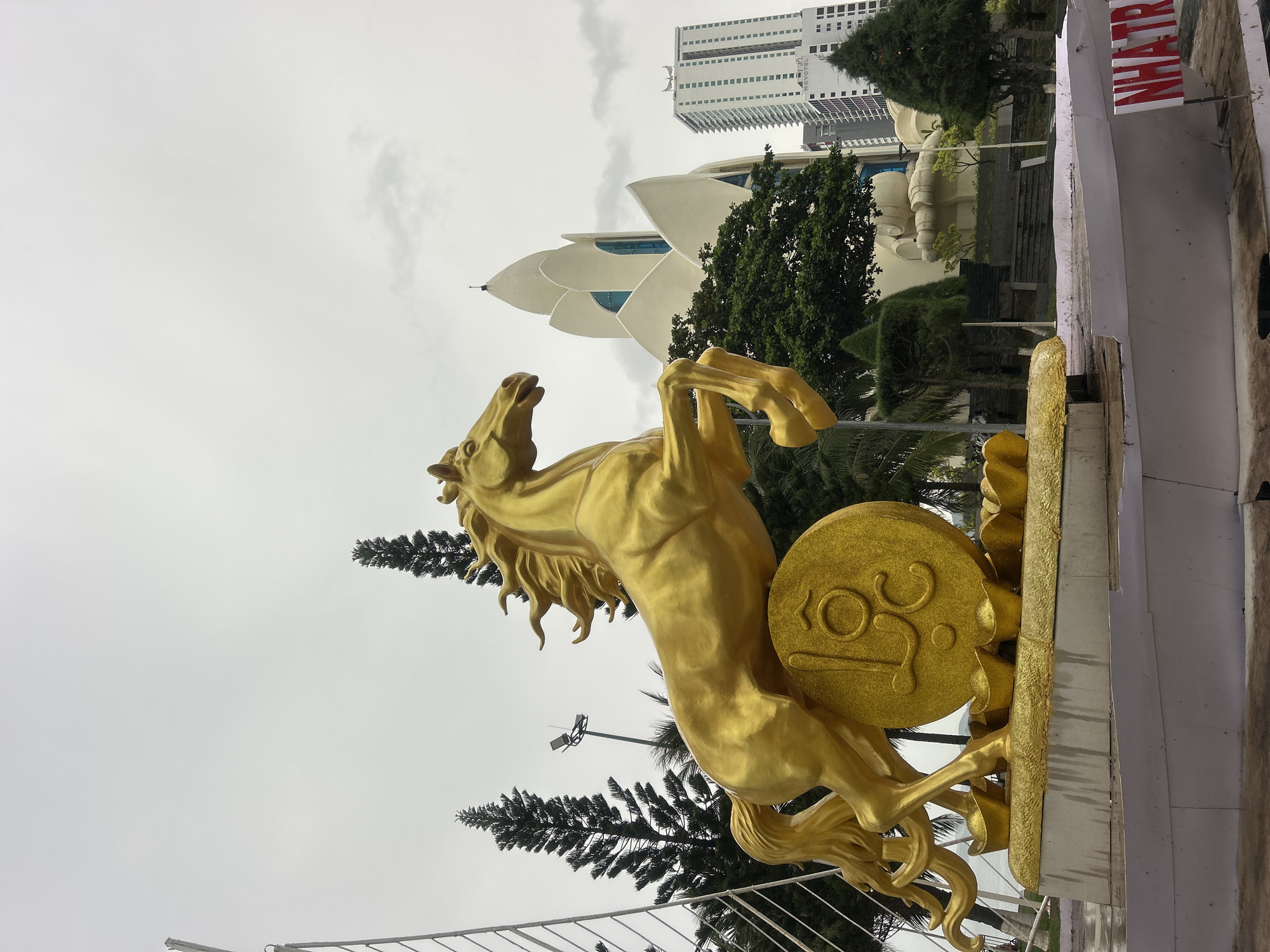 A golden horse statue is angled right in the center of the frame in Nha Trang Ward, Vietnam. It appears to be mounted on a platform with a golden disc that has decorative designs on it. The background features a building with a lotus-like design in white, a taller building in the distance, and trees. The sky is overcast, and there are other elements like lampposts and fencing visible as well. A sign that says NHATRA appears in the upper right corner.