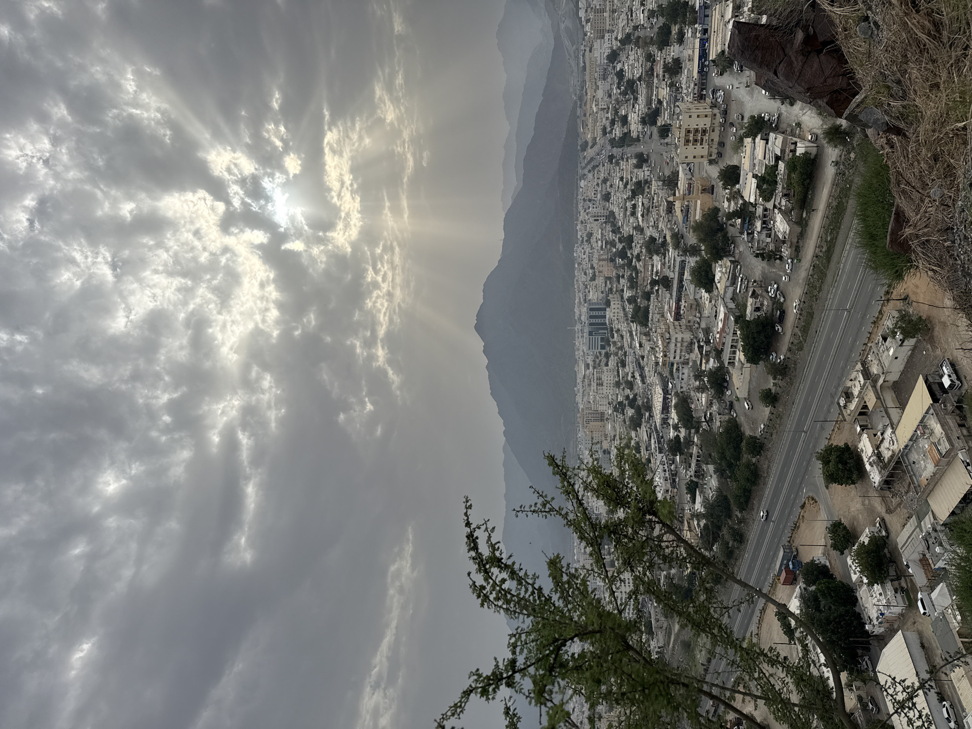 The image is a high-angle, vertical-oriented shot of Khor Fakkan, United Arab Emirates, taken on an overcast day with sun rays piercing through the clouds. Mountains form a backdrop to the cityscape. A road runs along the edge of the city, with buildings densely packed together. Vegetation is visible in the foreground and interspersed within the urban area. The shot seems to be taken from a vantage point on a slope or hill overlooking the city.
