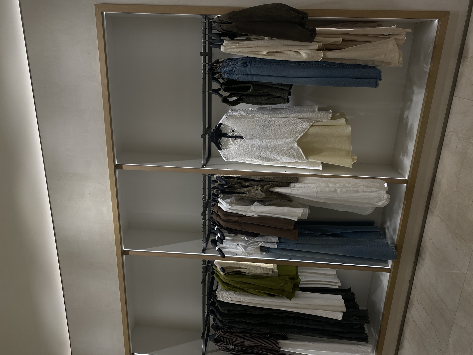 The image depicts clothing items arranged on shelves inside a store, likely a retail environment. The items on the top shelf include folded jackets, blouses, and blue jeans. The middle shelf features a white shirt, what appear to be jackets, and blue jeans. The lowest shelf contains folded clothes in green, black, and tan colors. These shelves are part of a store with neutral-toned walls. The exact location in Unknown, Unknown cannot be determined from the image.