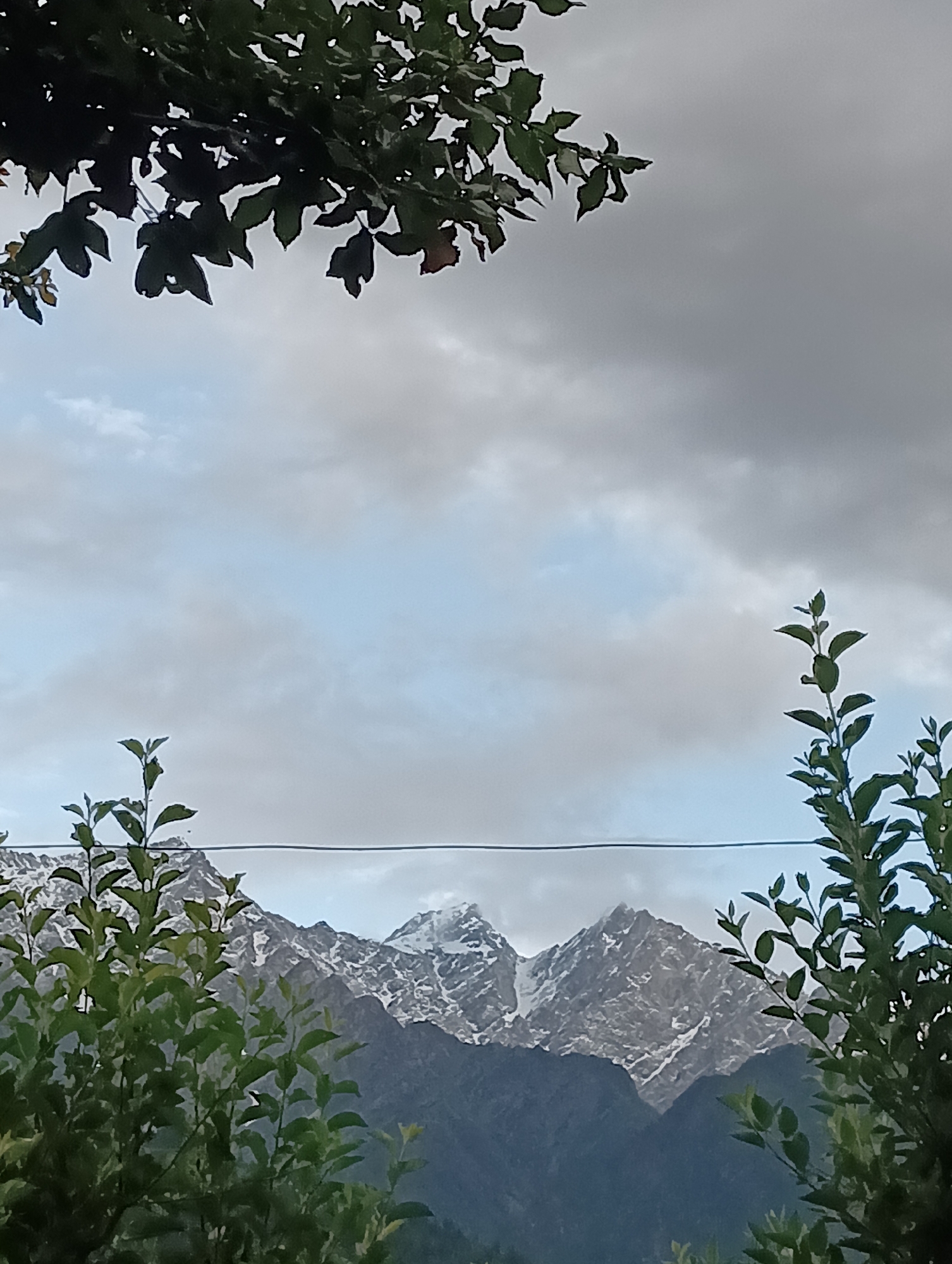 A photograph taken from Vashisht, India, captures a dramatic sky over snow-capped mountains. In the foreground, lush green foliage frames the scene, with dark leaves from a tree at the top and lighter, more delicate branches of smaller plants on the sides and bottom. A thin, dark wire stretches horizontally across the middle of the frame, dividing the sky and the mountainous landscape. The sky itself is a gradient of light blue and gray, with clouds creating a textured, somewhat overcast atmosphere. The mountains in the background are rugged and imposing, with distinct peaks, some covered in patches of white snow, contrasting with the darker, rocky slopes. The overall impression is one of natural beauty and dramatic weather, with a hint of human presence suggested by the wire.